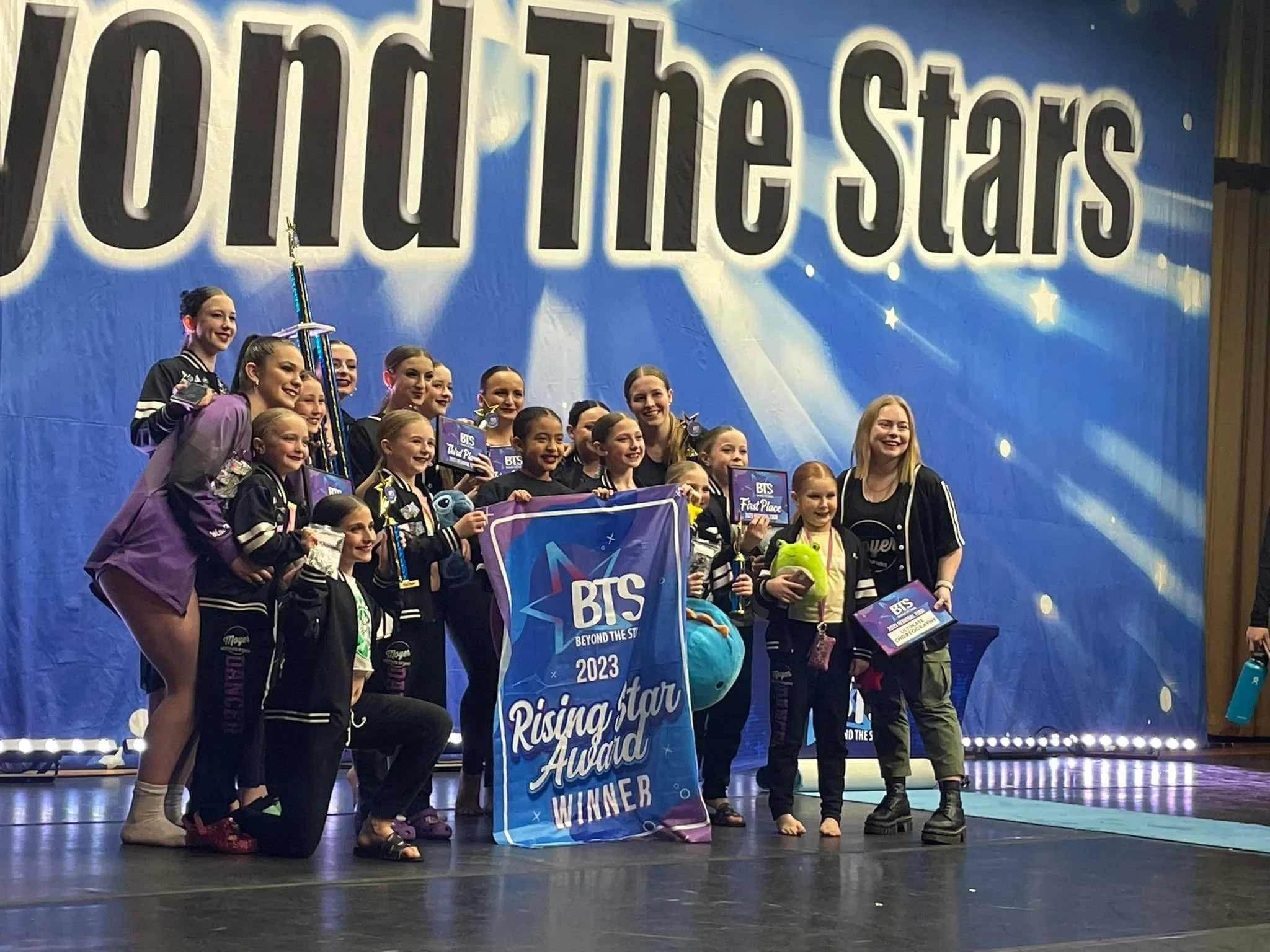 A group of young girls and their dance instructor showcase awards and a large trophy on stage at a dance competition called 'Beyond the Stars 2023.' They hold a banner reading 'Rising Star Award Winner' and various prizes, smiling happily.