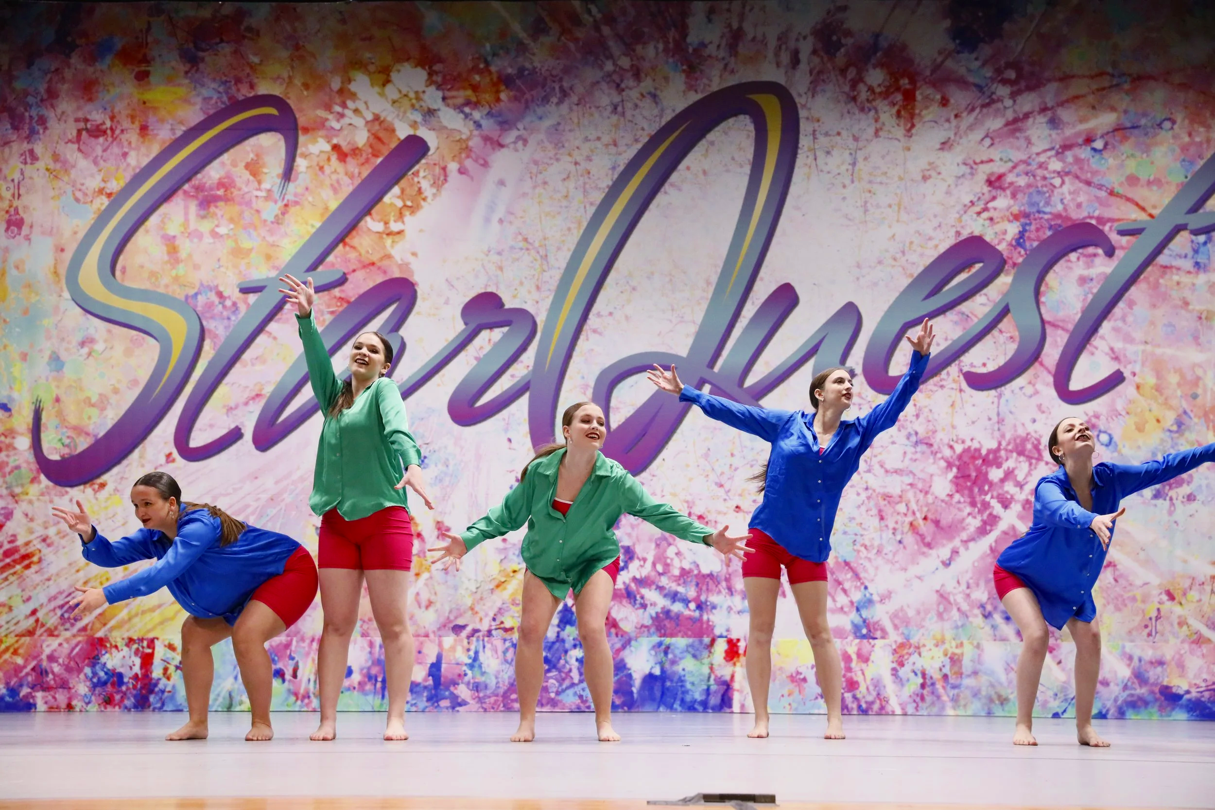 Five young women performing a dance on stage with a colorful abstract background and the word 'Stardust' in large cursive letters.
