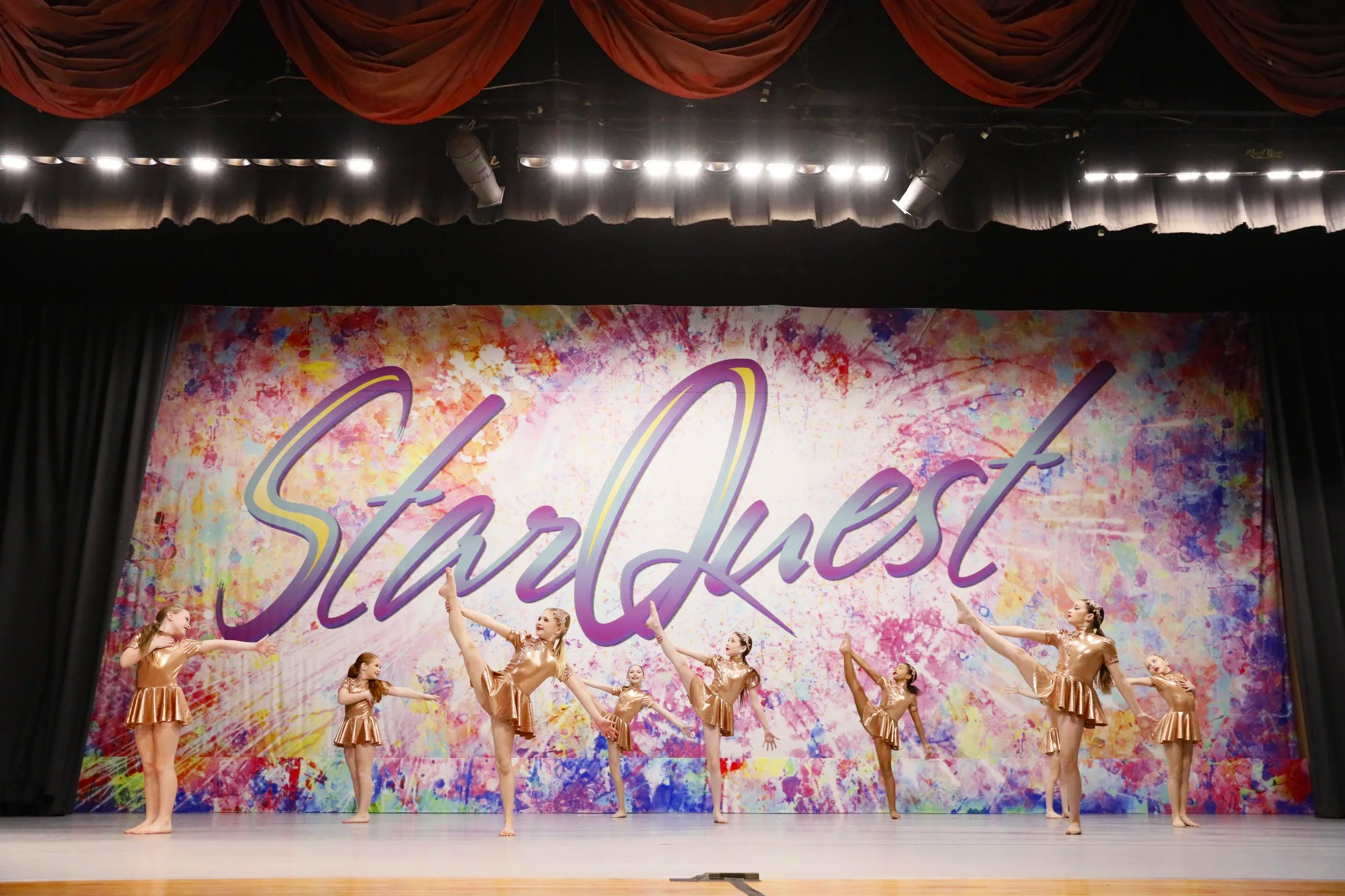 Group of young dancers in gold costumes performing a dance routine on stage with a colorful background reading 'Starquest'.