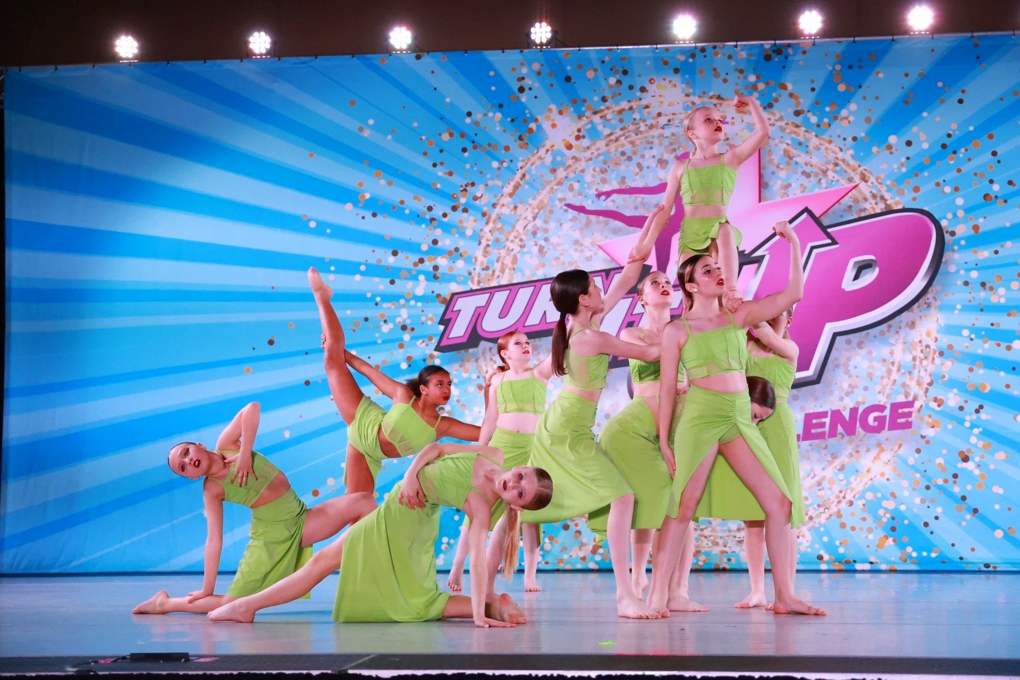 A group of young female dancers in matching green costumes perform a dance routine on stage, with a bright blue backdrop featuring a logo and colorful design elements.