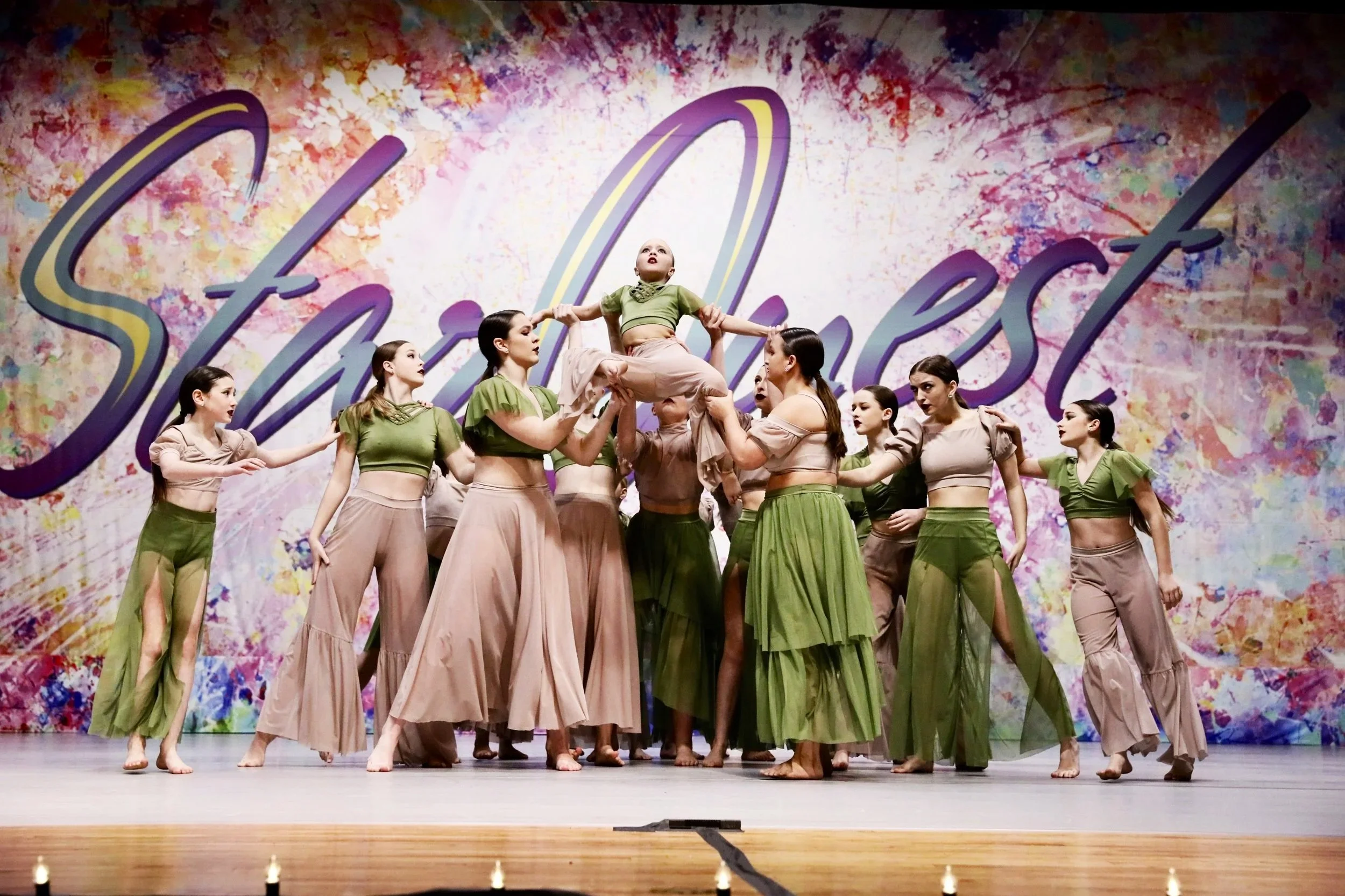 A group of female dancers performing on stage, holding a young girl above their heads, with a colorful backdrop that says 'Starquest'.