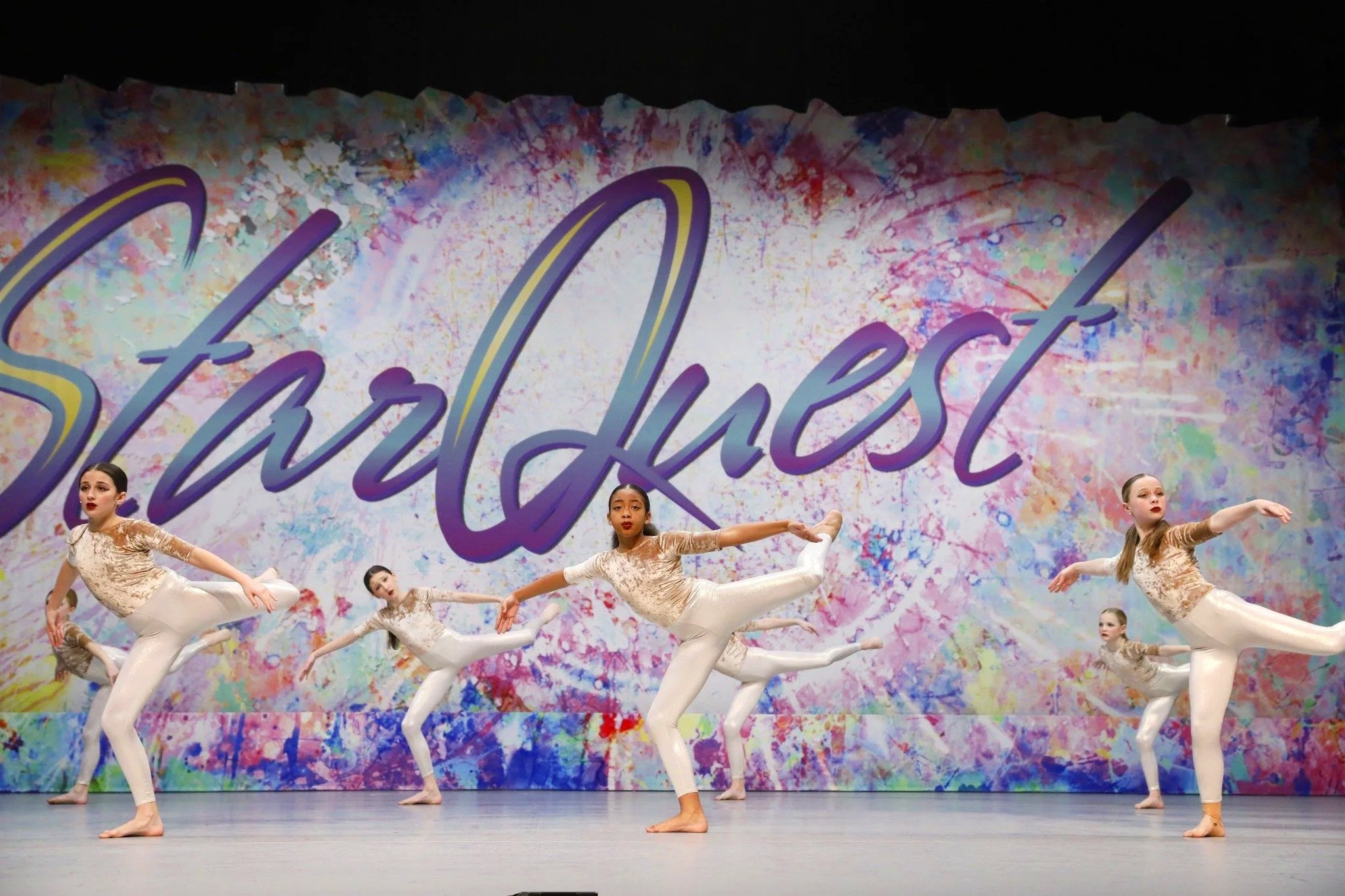 Dancers perform on stage in front of a colorful backdrop with the words 'Star Quest' written on it.