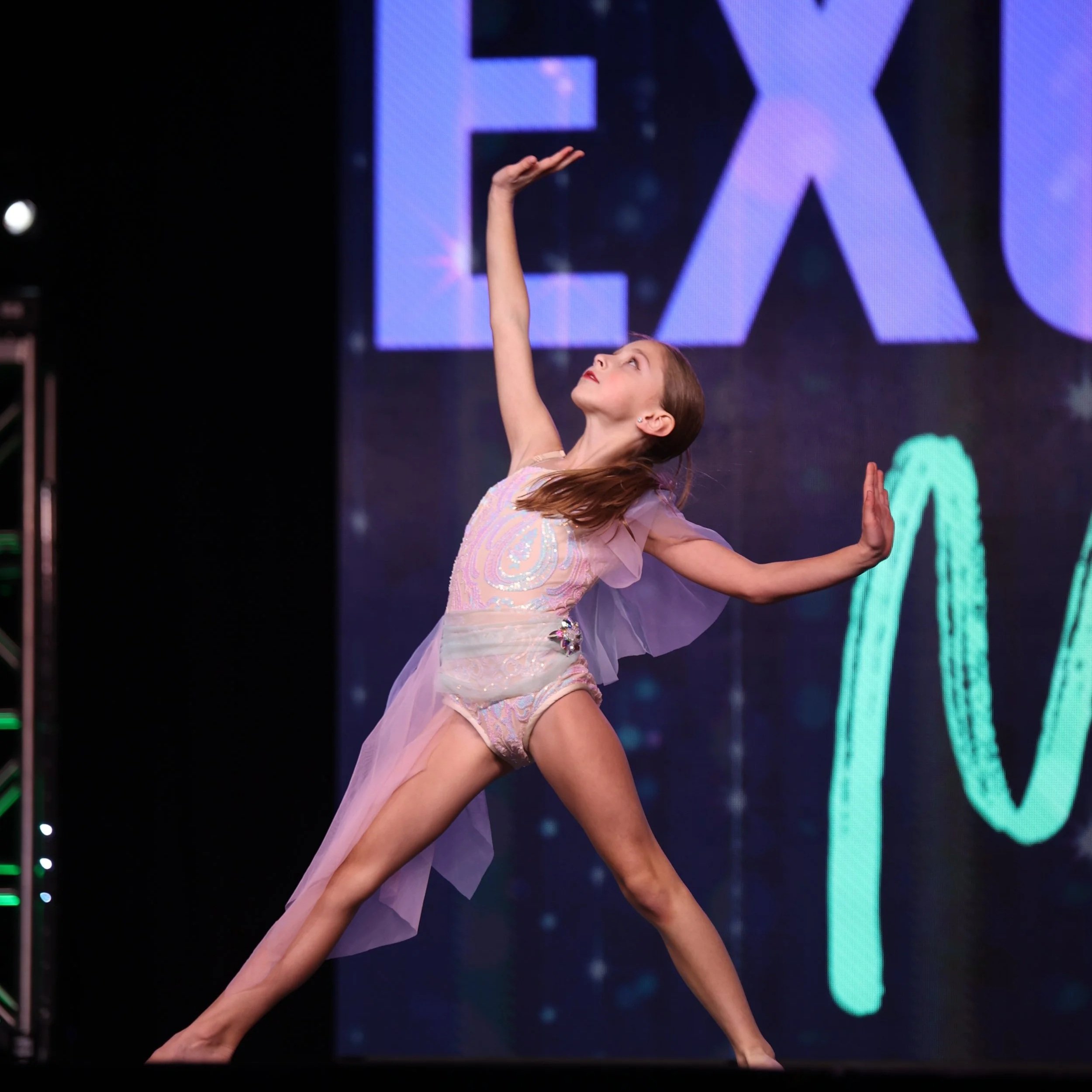 A dancer performing on stage in a sparkling costume, with large illuminated letters in the background.
