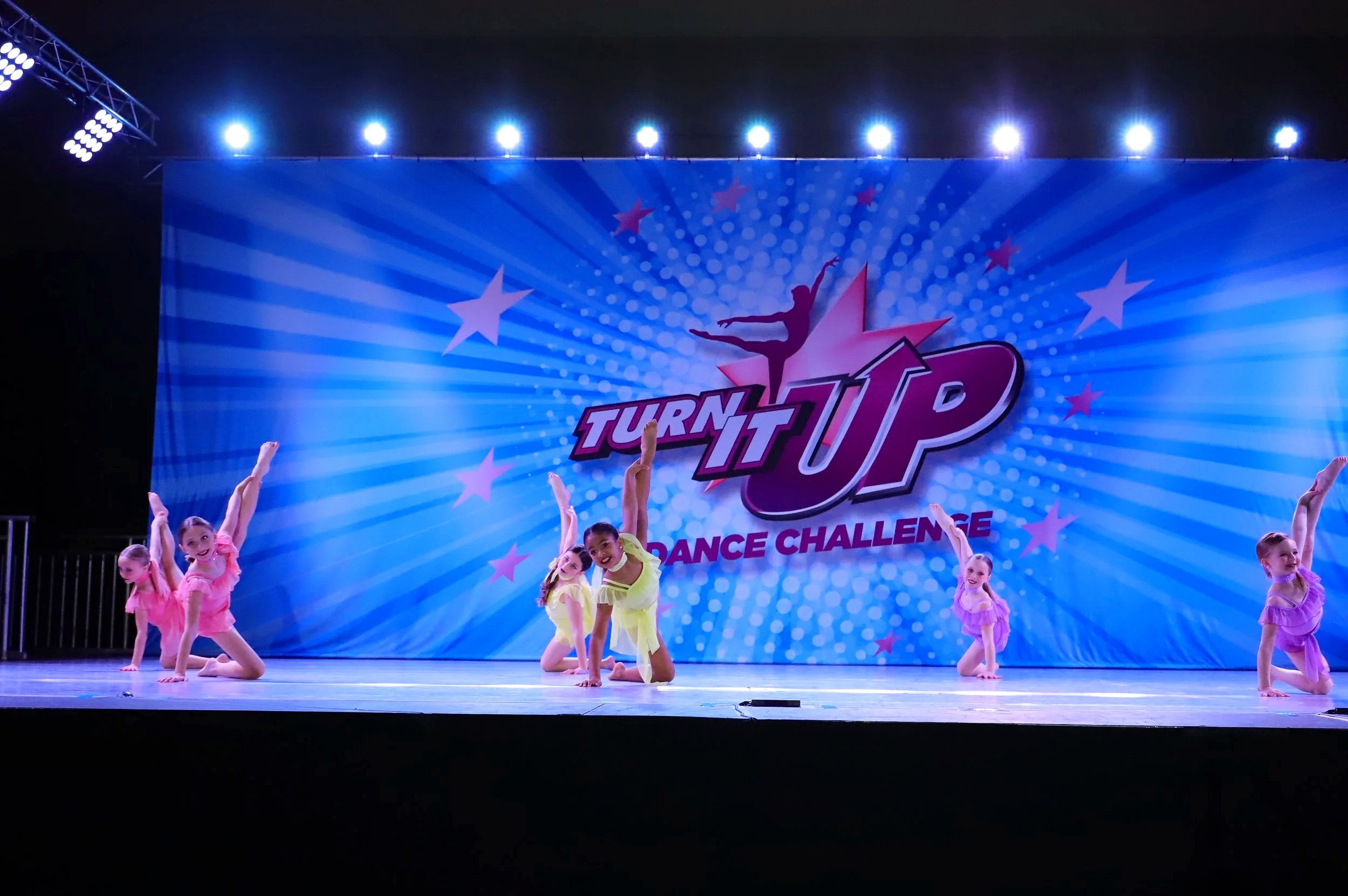 Young girls dressed in colorful costumes performing a dance on stage at a dance challenge event with a bright blue backdrop and a large logo reading 'Turn It Up'.