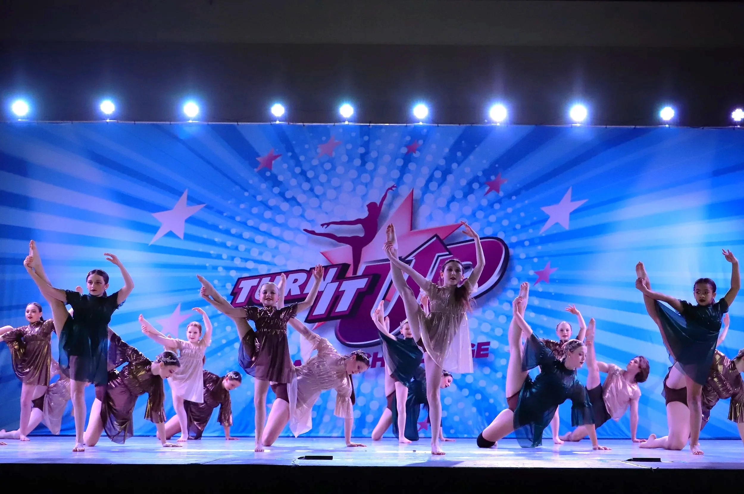Group of young dancers performing a ballet routine on stage, with a blue background featuring stars and the text 'Turn It Up'.