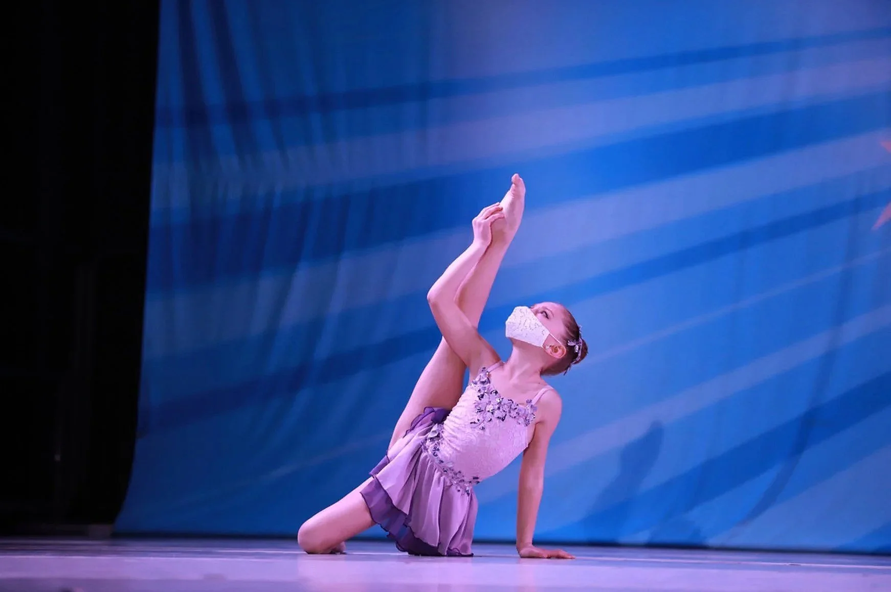 A young girl in a purple dance costume performs a routine on stage, balancing on one hand with her legs extended upwards, wearing a face mask.