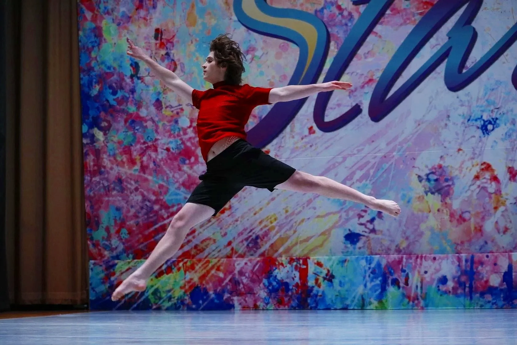 A male dancer in a red shirt and black shorts performing a leap on a stage with a colorful, splattered paint background.