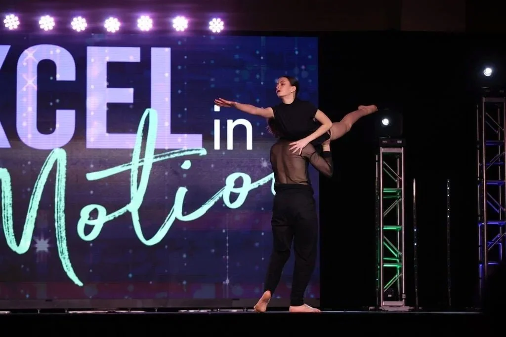 Two performers doing a dance routine on stage, one lifting the other while balancing on one foot. The stage features a large screen in the background with the words "Dance in Motion" and colorful lighting.