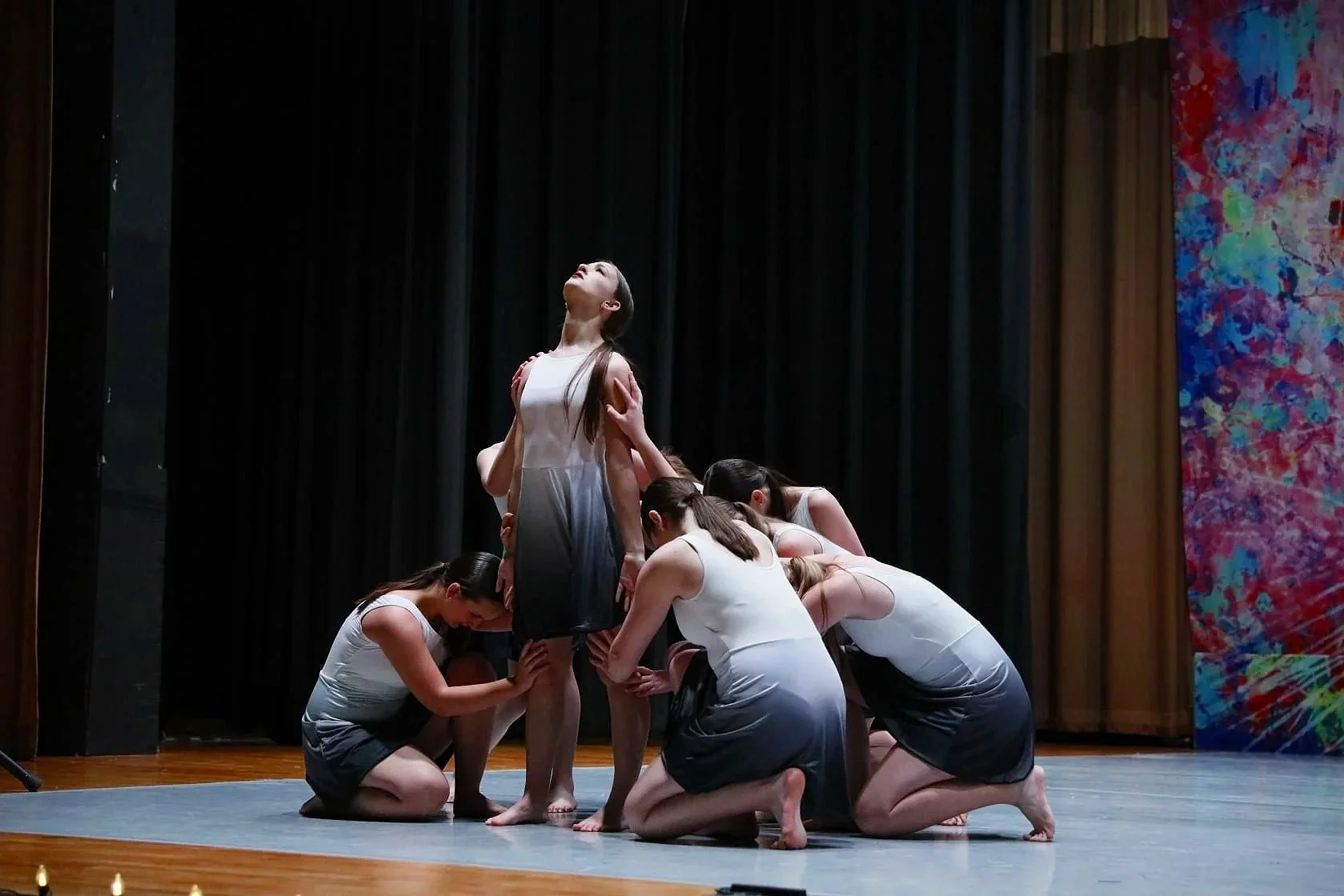 A dance performance on stage with a woman in the center surrounded by several women supporting her, some kneeling and others bending over, all wearing white dresses. The woman in the center looks upward, with a dark curtain behind them and colorful a