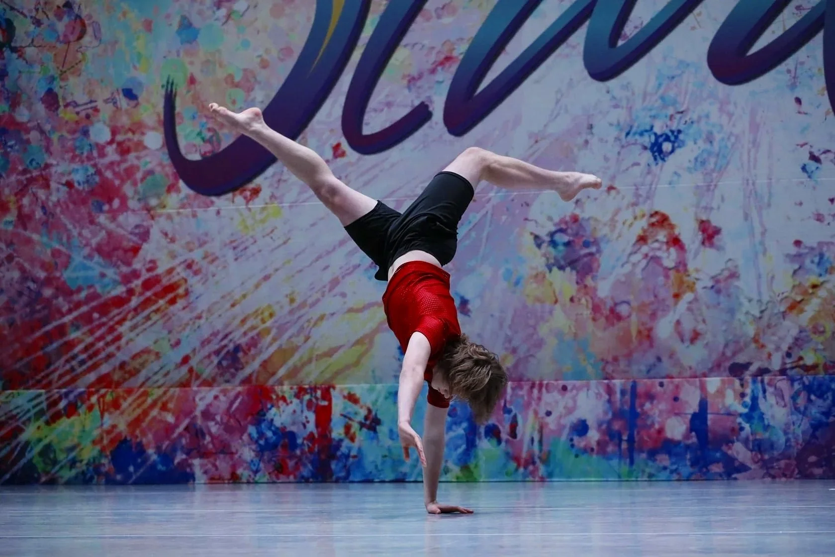 A person performing a handstand or acrobat on stage with a colorful, abstract background.