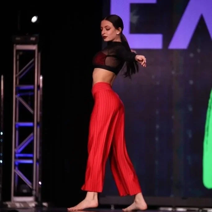 A woman dancing on stage, wearing a black sheer long sleeve crop top and high-waisted red pants with vertical stripes.