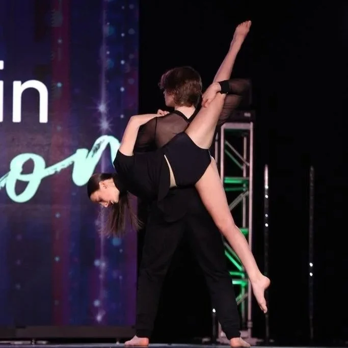 Two women performing a dance lift on stage with a backdrop displaying lights and text.