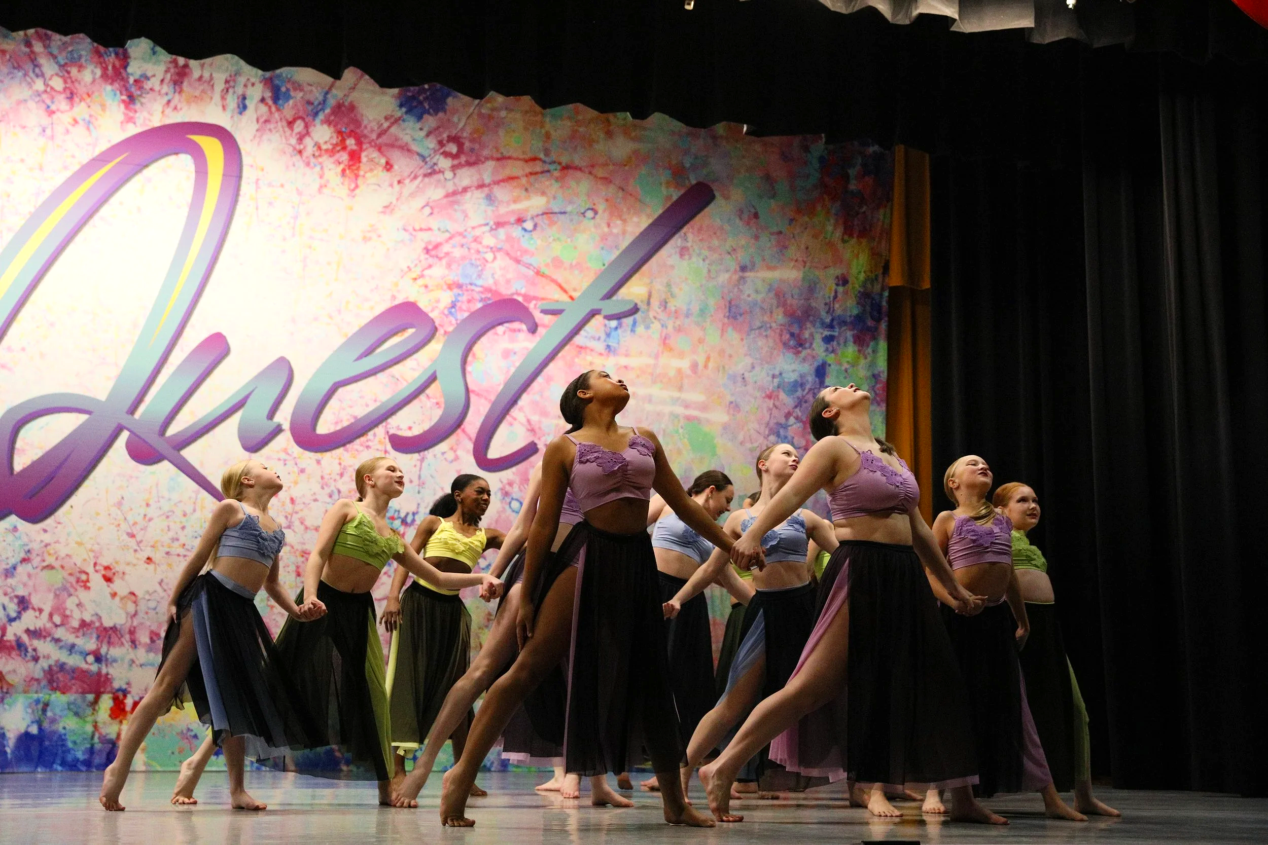 Group of dancers on stage performing with hands linked, wearing pastel-colored crop tops and long skirts, with a colorful backdrop displaying the word 'Duest' in stylized script.