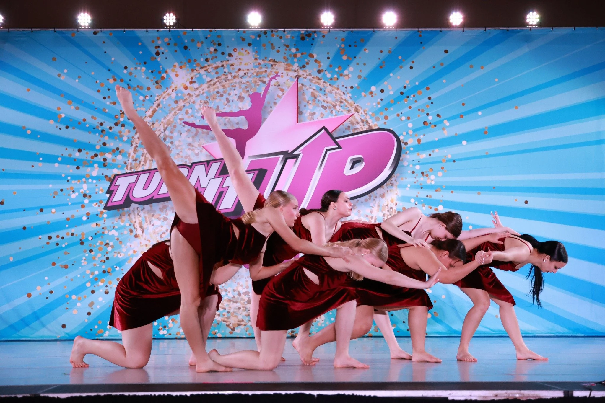A group of six female dancers in red dresses performing a synchronized dance move on stage with a background that says 'Turn Up' and features a dancer silhouette and a star design.