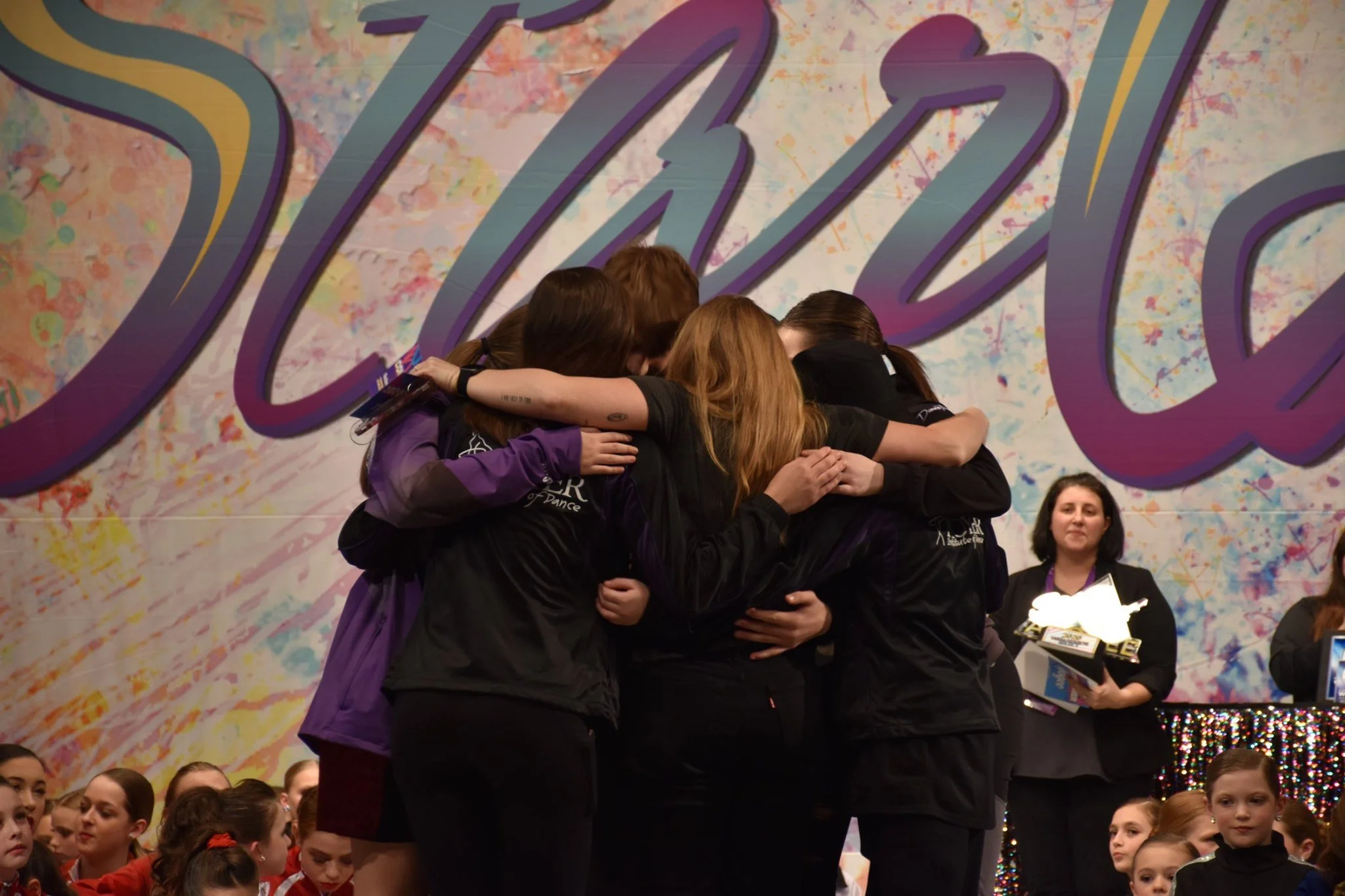 Group of young girls in black jackets hugging each other in a circle on stage, with a large colorful background and other girls and women watching.