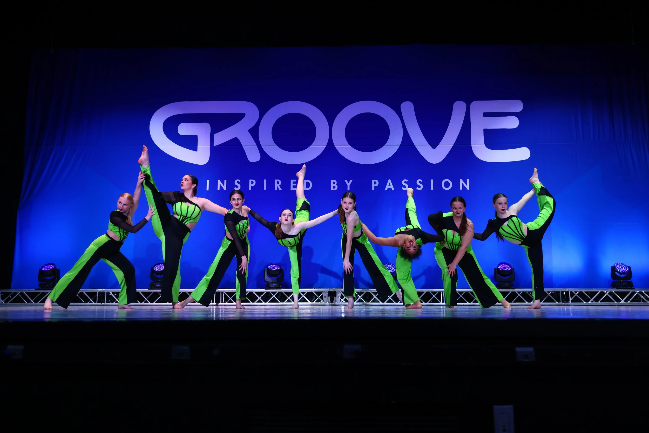 A group of eight young female dancers in matching neon green and black costumes perform on stage in front of a blue backdrop with the words 'GROOVE' and 'INSPIRED BY PASSION'.
