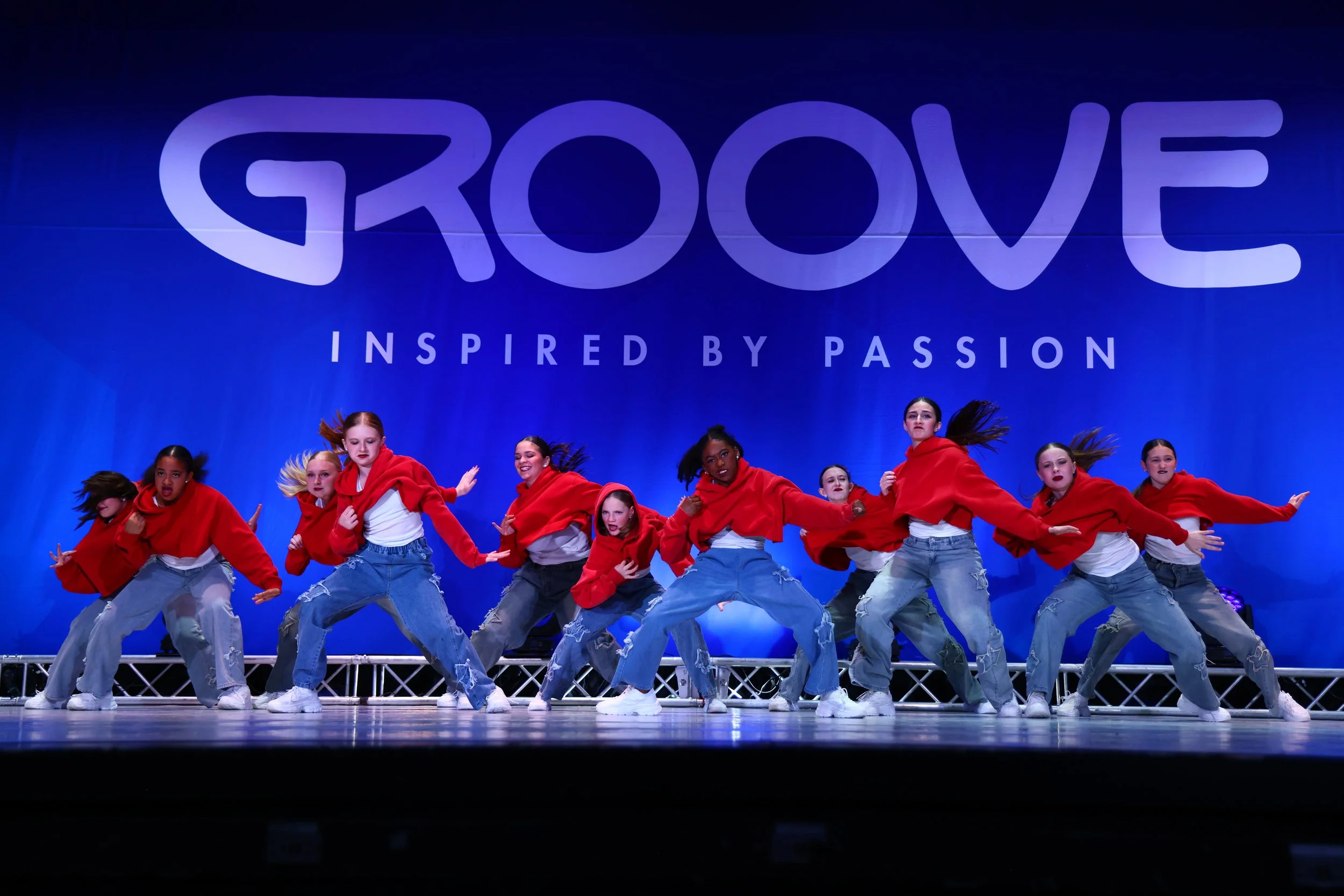 A group of young female dancers performing a hip hop dance routine on stage, wearing red hoodies, white T-shirts, baggy ripped jeans, and sneakers, with a blue backdrop that reads 'GROOVE inspired by passion'.