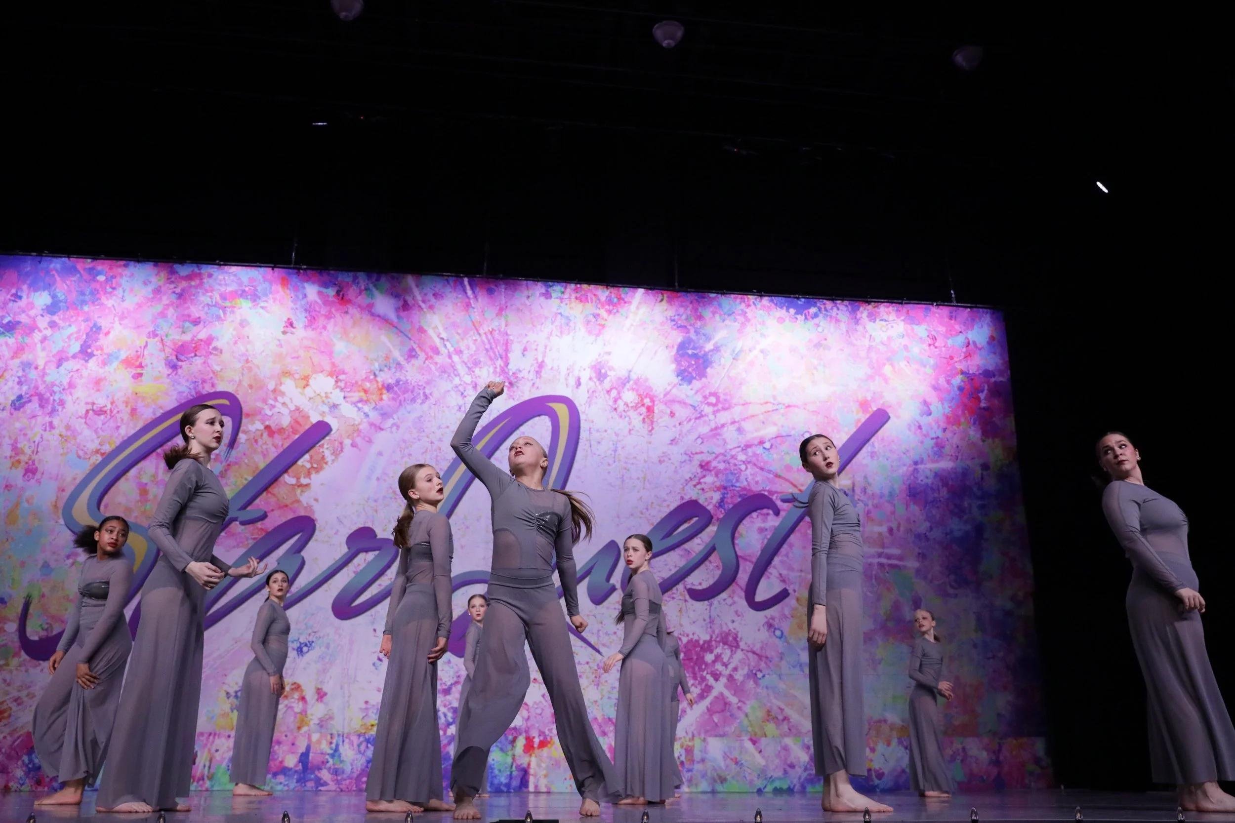 A group of nine young girls performing a dance on stage, dressed in matching long gray dresses. They are positioned in front of a colorful background with the word 'Dance' written in large cursive script.