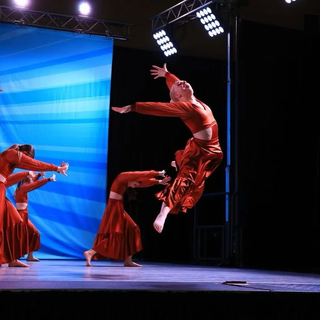 Dancer jumping in mid-air during a stage performance, with other dancers in matching costumes behind her.
