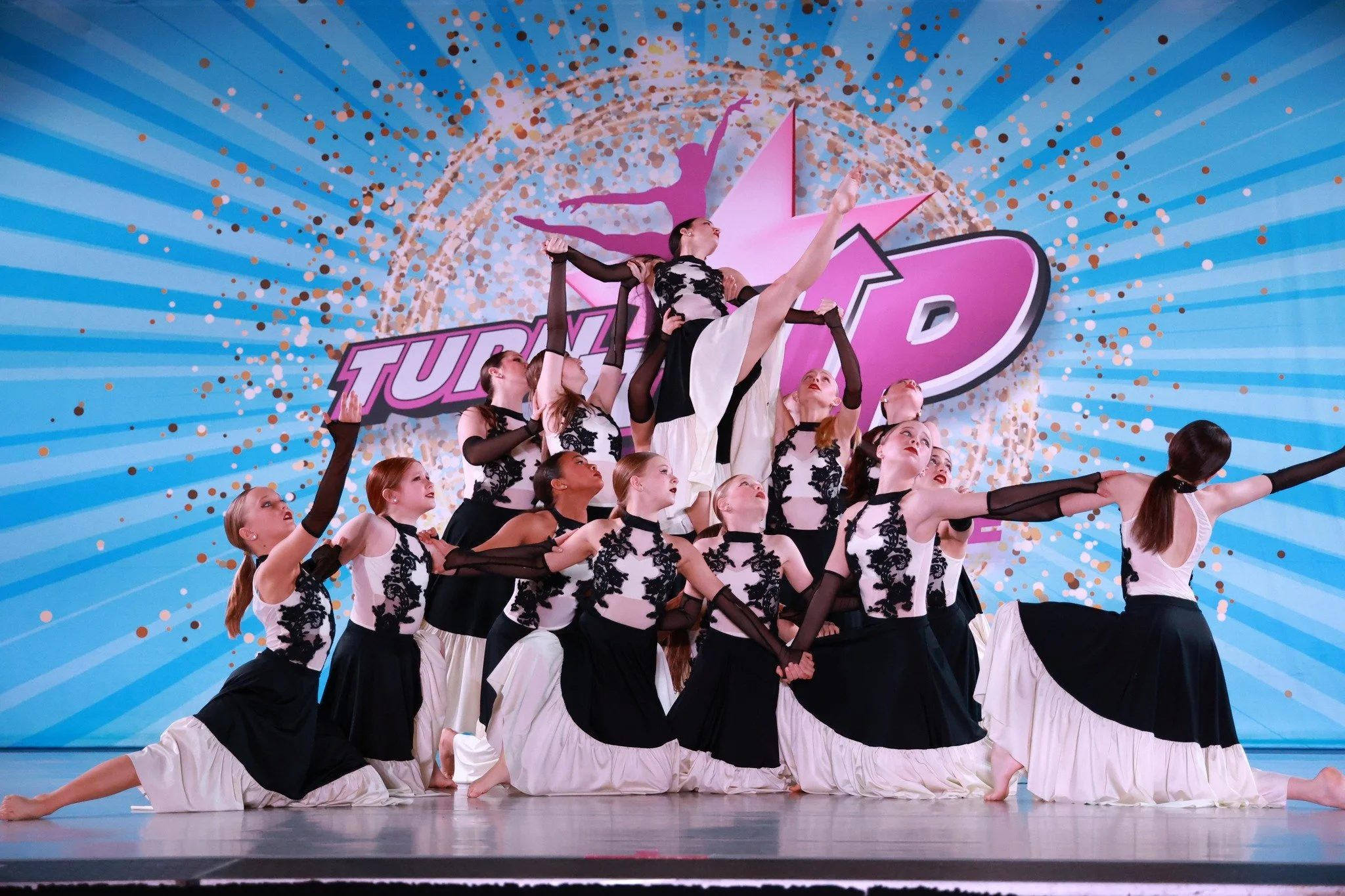 A dance team performing a choreographed lift on stage with a colorful backdrop that says 'Turn Up' and features a pink star. The dancers are dressed in coordinated black and white costumes with lace details and black gloves.