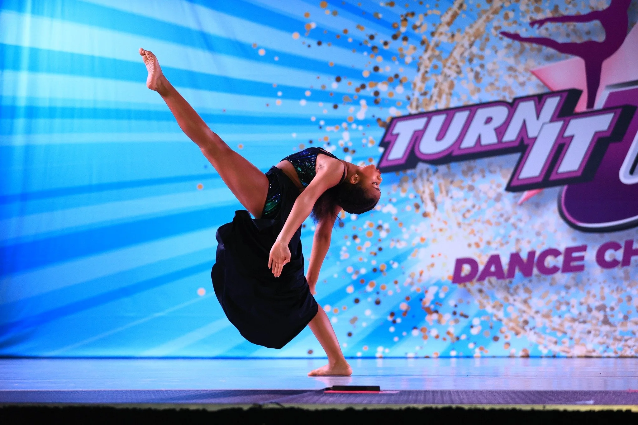 A dancer performing a handstand on stage during a dance competition, with a colorful background and the word 'Turn It' visible.