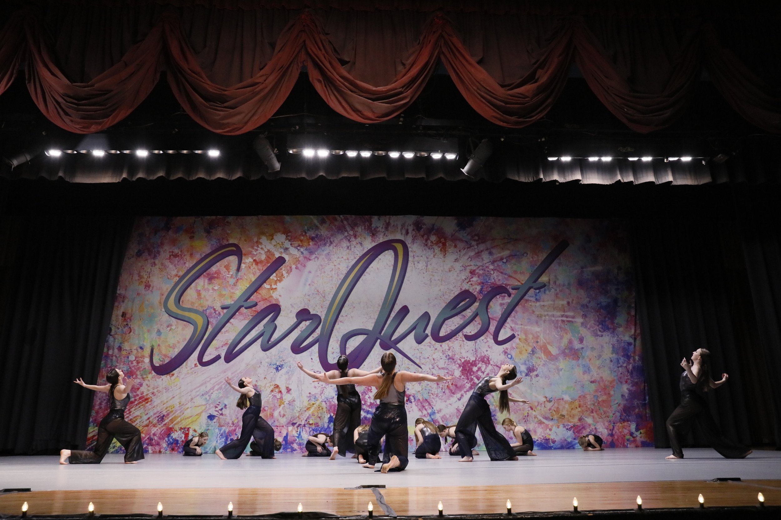 Dance performance on stage with young dancers in black costumes, performing in front of a colorful backdrop with the word 'StarQuest'.