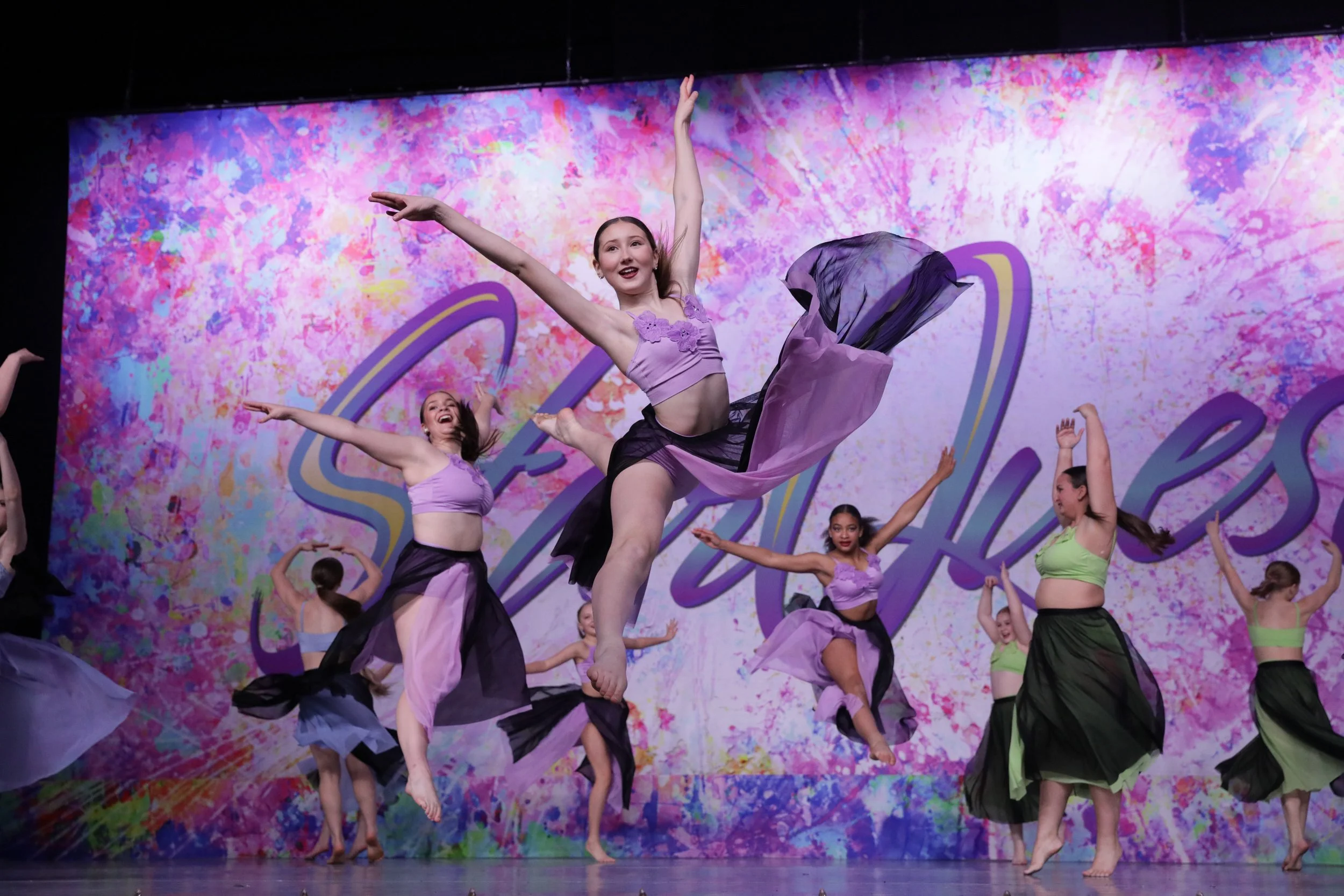 A group of young dancers performing a ballet routine on stage, with a colorful splatter paint backdrop featuring the word 'Sweet' in cursive.