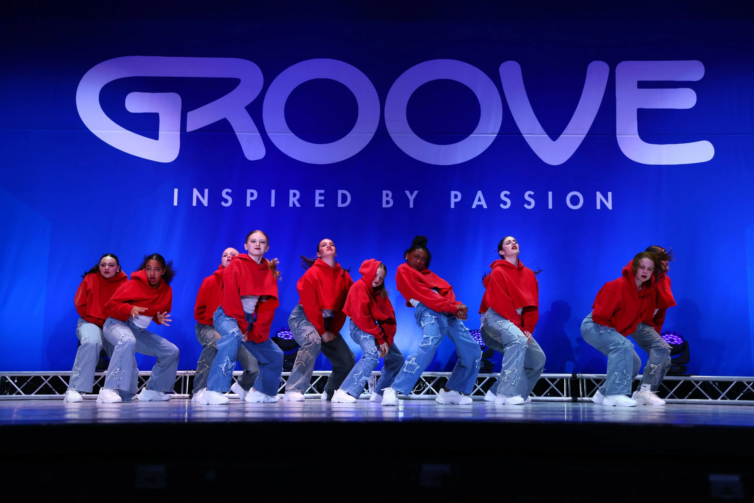 Group of young female dancers in red hoodies and ripped jeans performing on stage with a blue backdrop that reads 'GROOVE' and 'INSPIRIED BY PASSION'.