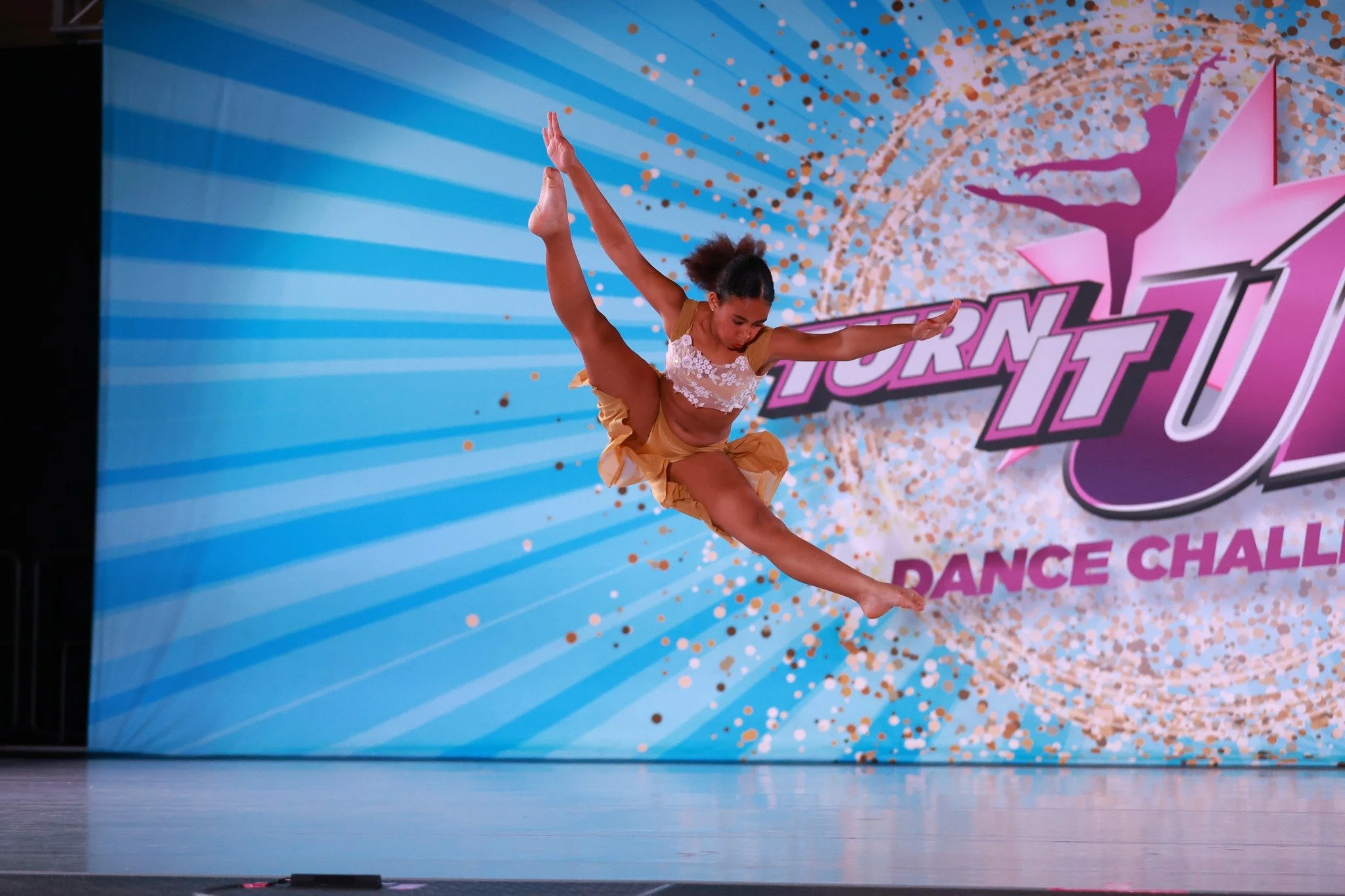 A young female dancer performing a leap on stage during a dance competition, with a colorful backdrop that reads 'Turn It Up Dance Challenge'.