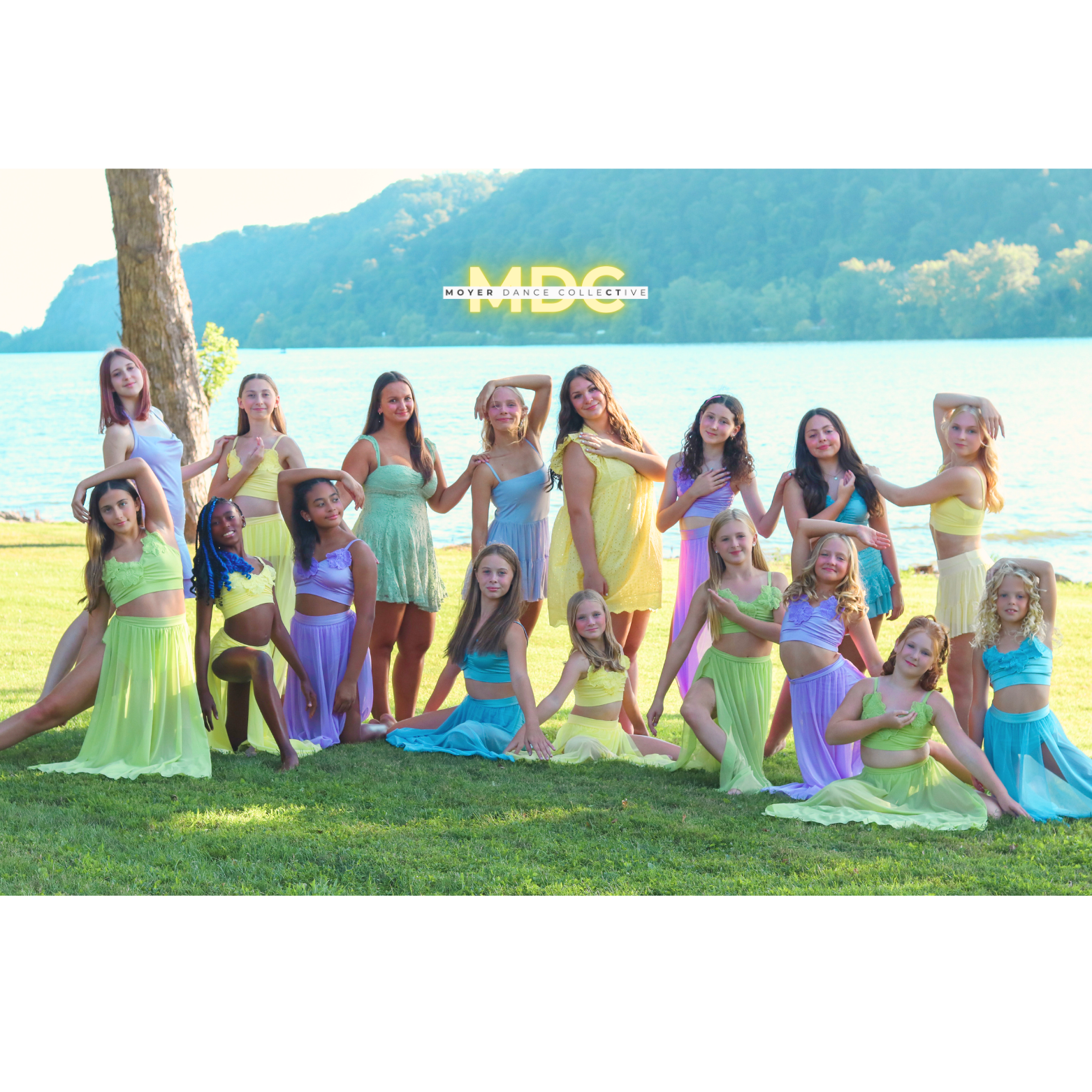 Group of young dancers in pastel-colored dresses posing outdoors by a river with mountains in the background, promoting Moyer Dance Collective.