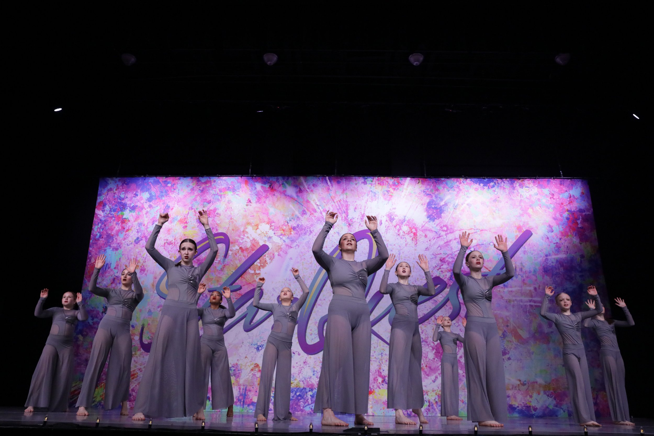 A group of female dancers in gray costumes performing on stage against a colorful, abstract backdrop.