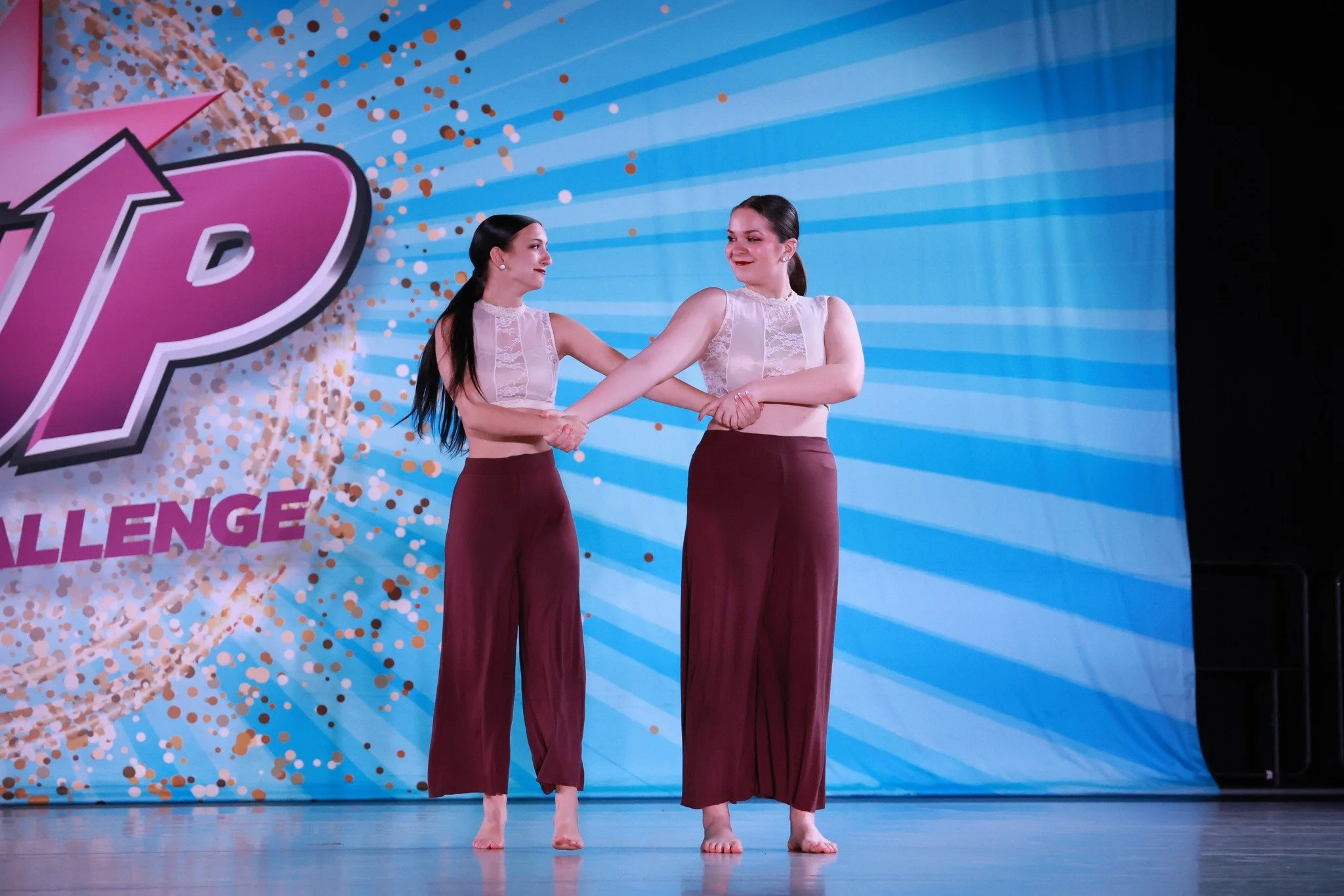 Two women performing a dance routine on stage, holding hands and smiling, with a blue and pink backdrop that reads 'CHALLENGE' and features a large arrow and confetti design.