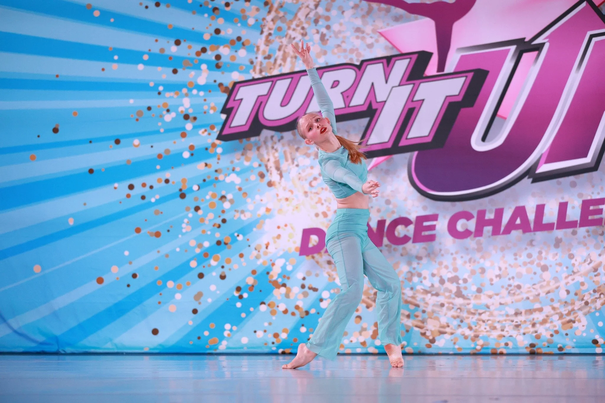 A young female dancer in a turquoise outfit performing a dance routine on stage with a backdrop that reads 'Turn Up Dance Challenge' and has confetti and bright colors.