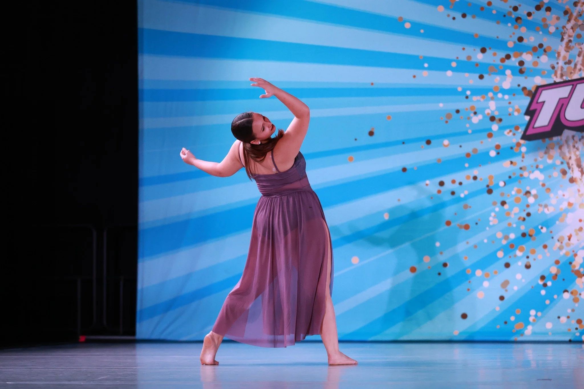 A woman in a purple dress dancing on stage with a colorful, sparkly background.