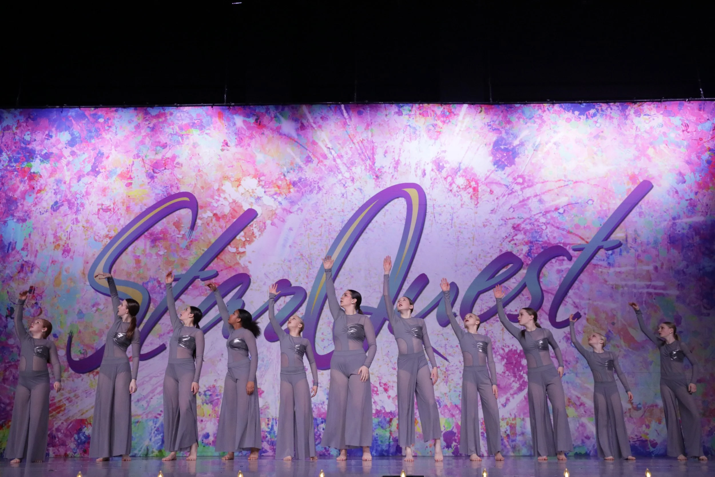 Group of young women in matching gray costumes performing a dance on stage with a colorful backdrop that says 'Skyfest'.