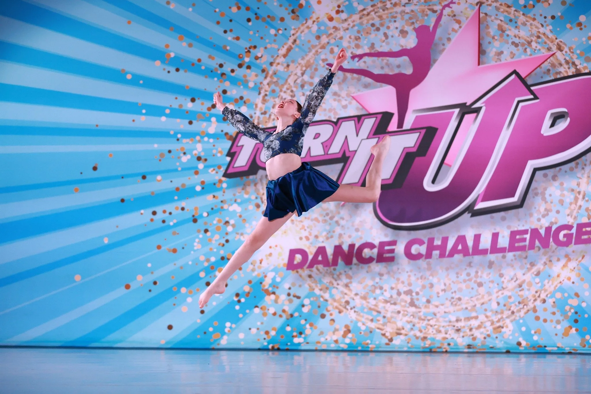 A female dancer jumps in mid-air during a dance competition, with a colorful backdrop displaying the words 'Turn It Up Dance Challenge' and a star logo.
