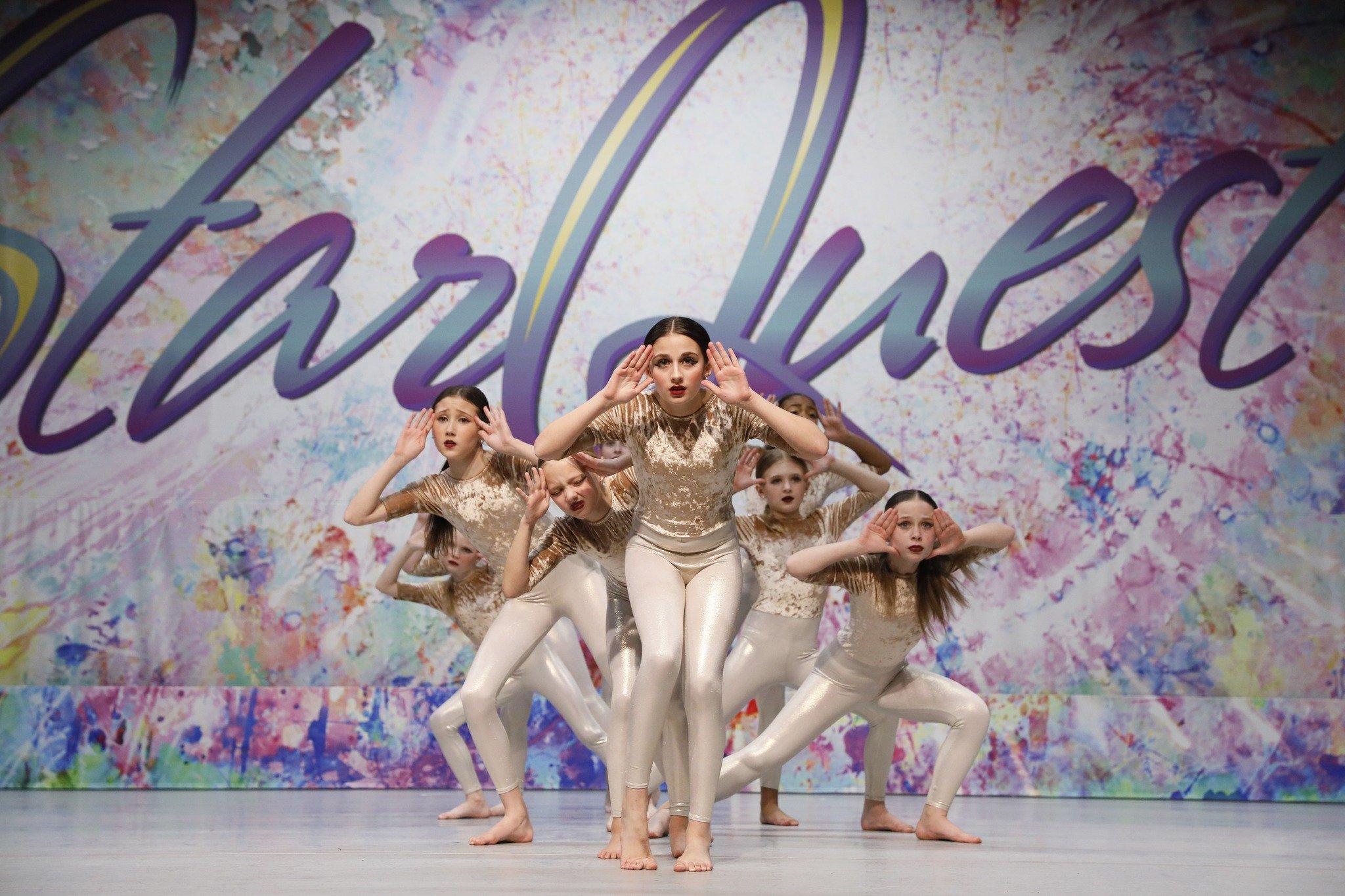Group of young girls performing a dance on stage in matching beige and gold costumes, with a colorful splashed background and large text saying 'Allieluiel'.