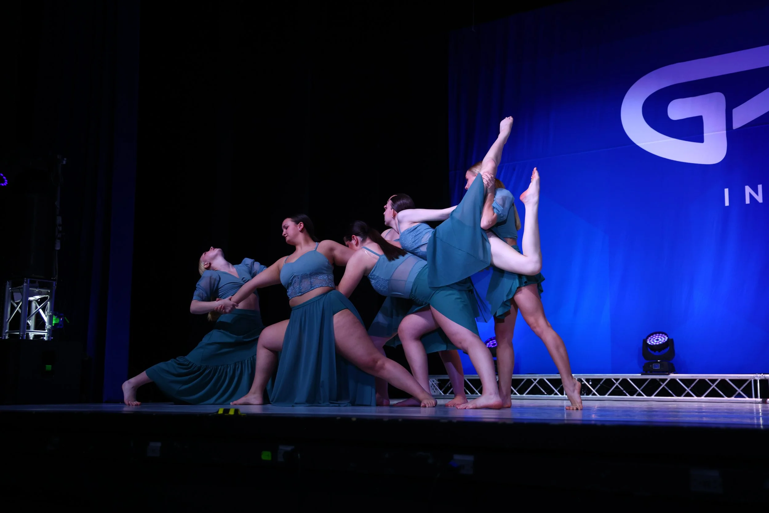 Group of five female dancers performing a contemporary dance on stage, wearing matching teal costumes, with a blue backdrop and stage lights.