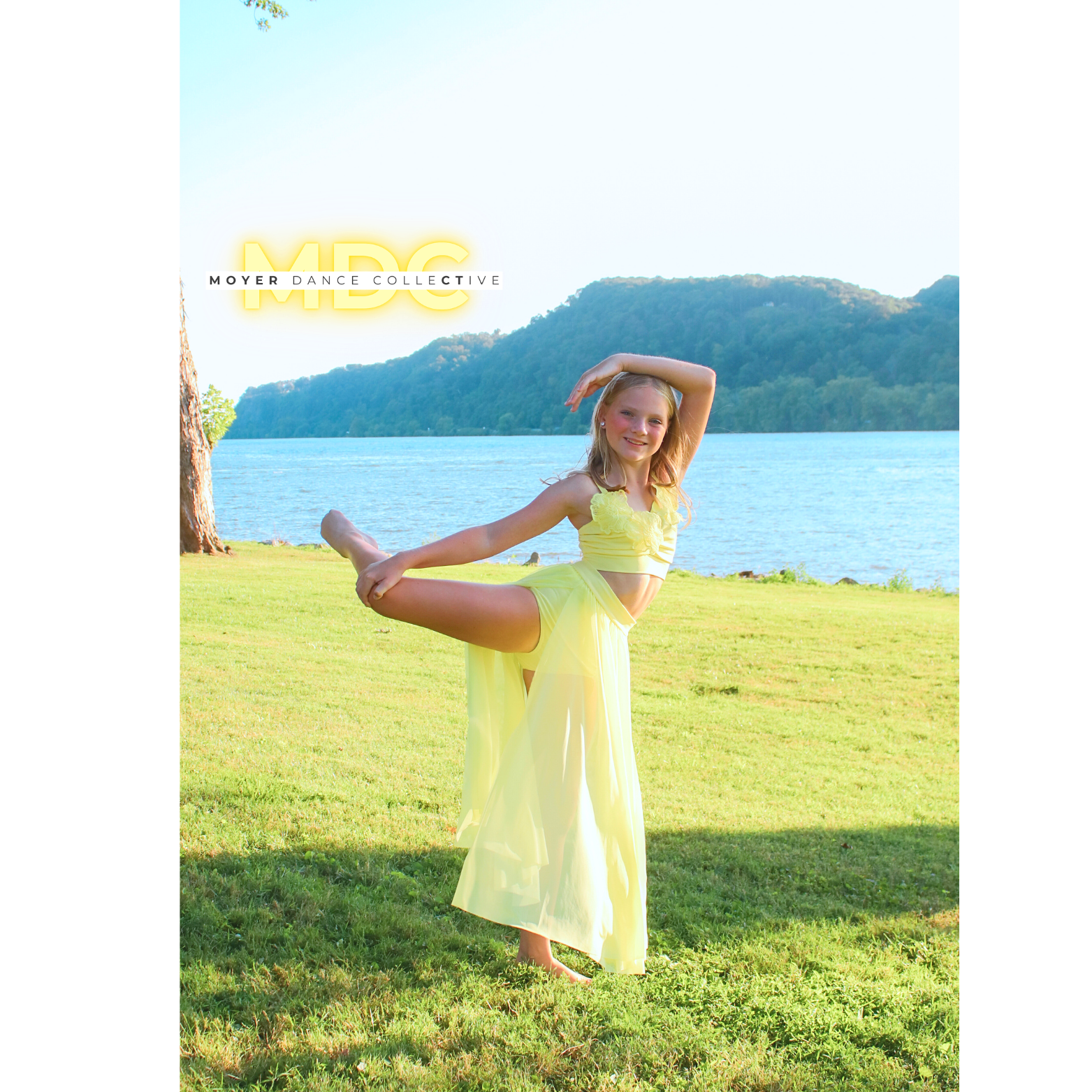A young dancer in a yellow dance costume doing a pose outdoors by a river with hills in the background.