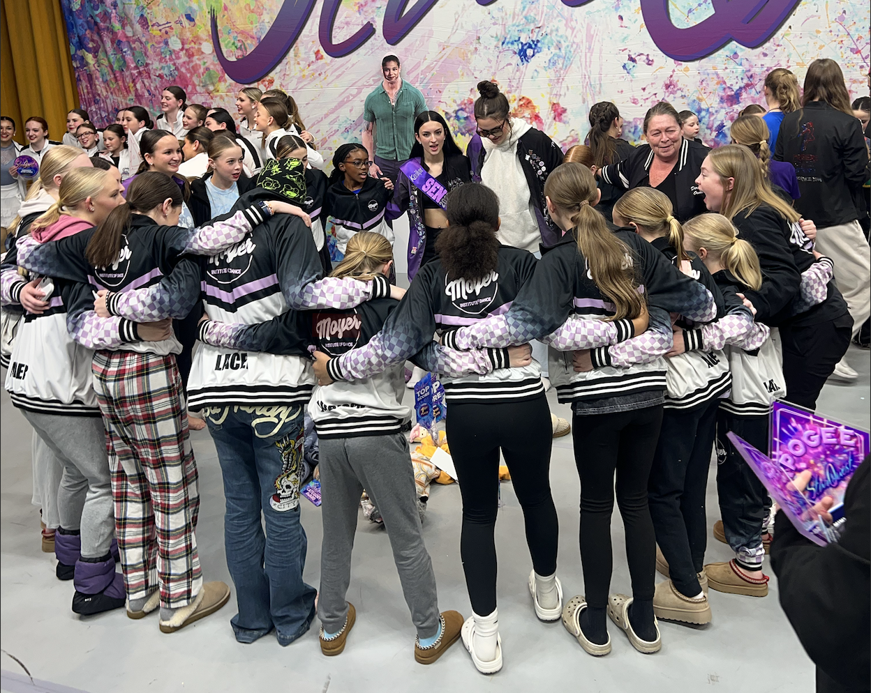 Group of young girls and women in matching jackets and casual clothing, embracing in a circle at a dance competition or event, with a colorful painted backdrop.