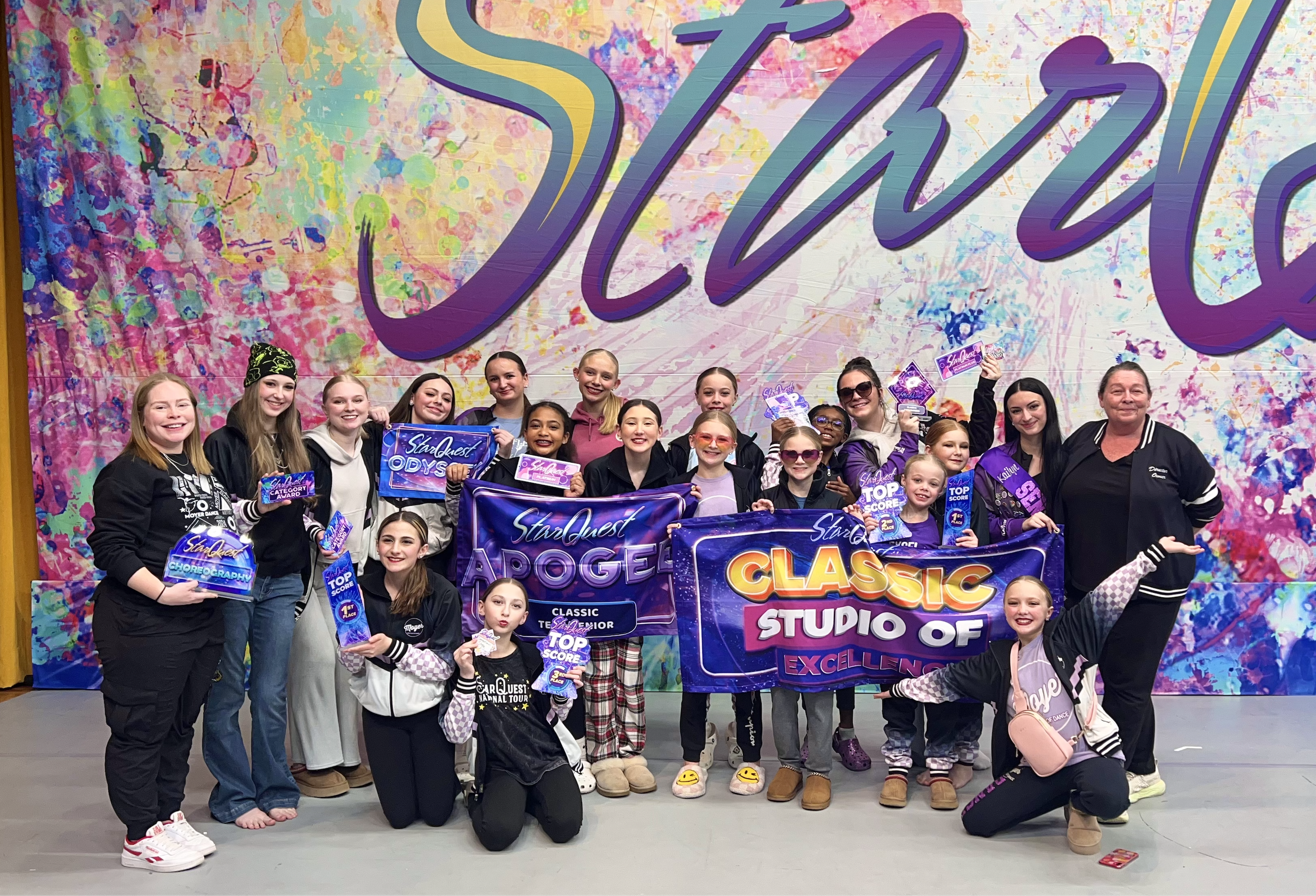 Group of young girls and their coach posing with awards on a stage in front of a colorful, artistic backdrop that says 'Studio.' The girls are holding banners and certificates with phrases like 'StarQuest Apollo,' 'Classic,' 'Studio of Excellence,' a