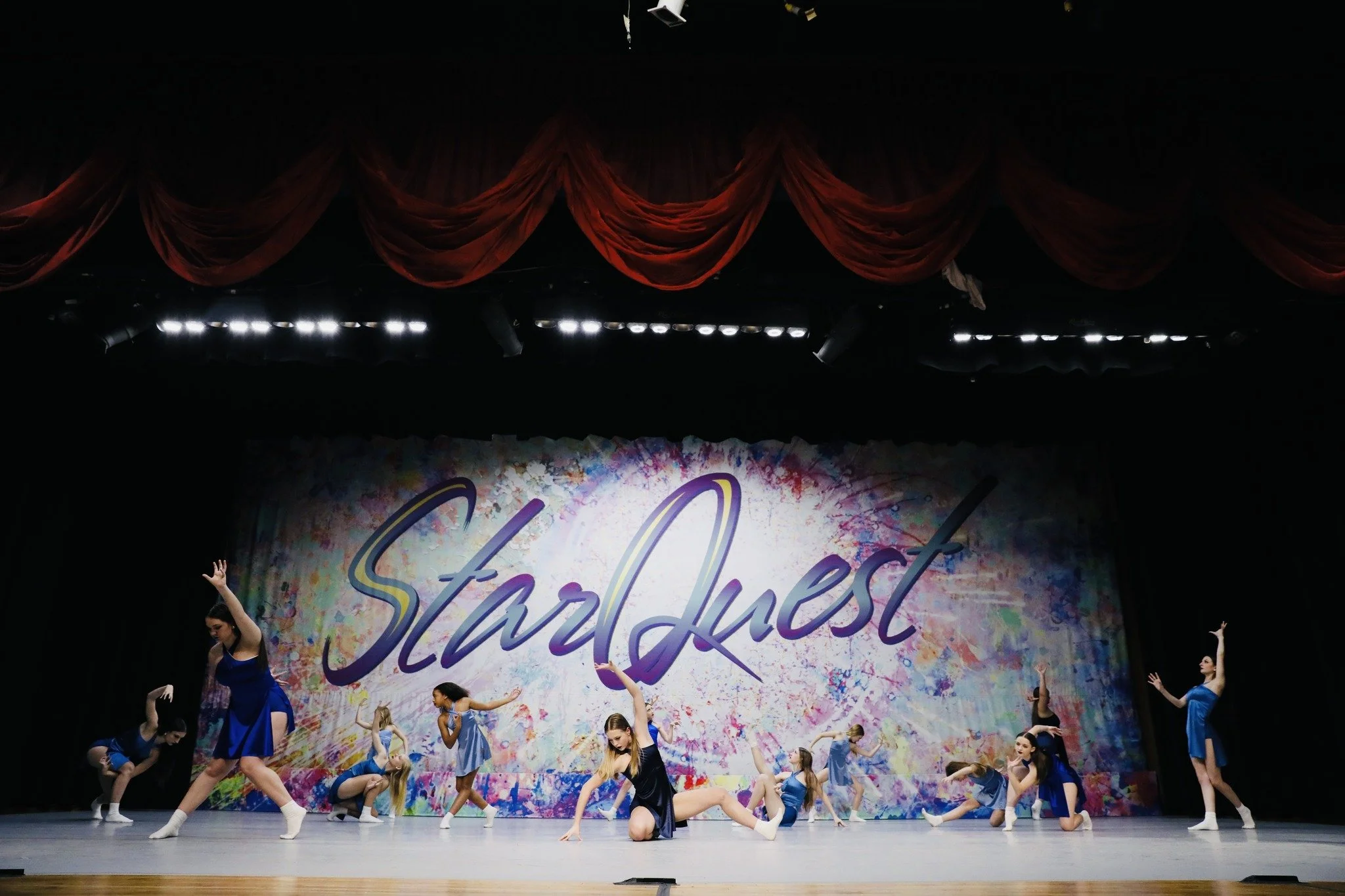 Dance performance on stage with ten young girls in blue costumes, some kneeling, some standing with arms raised, in front of a colorful backdrop with the words 'StarQuest'