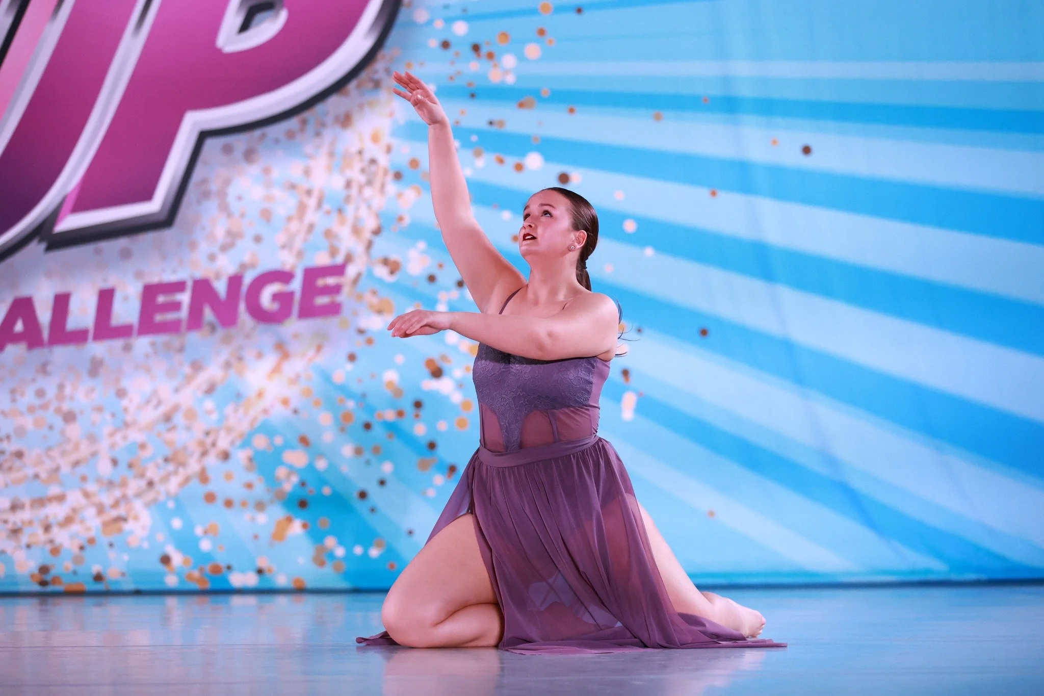 A young female ballet dancer performing on stage with a blue background and confetti, wearing a purple dance costume, kneeling on the floor with her arm extended upward.
