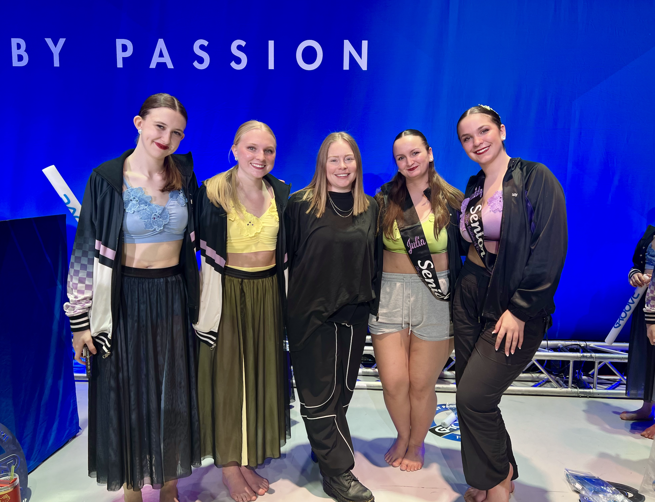 Group of five women standing together, smiling on a stage with a blue background. They are dressed in casual and athletic gear, some wearing black jackets and colorful tops, with one woman in the center wearing black pants and the others in shorts or