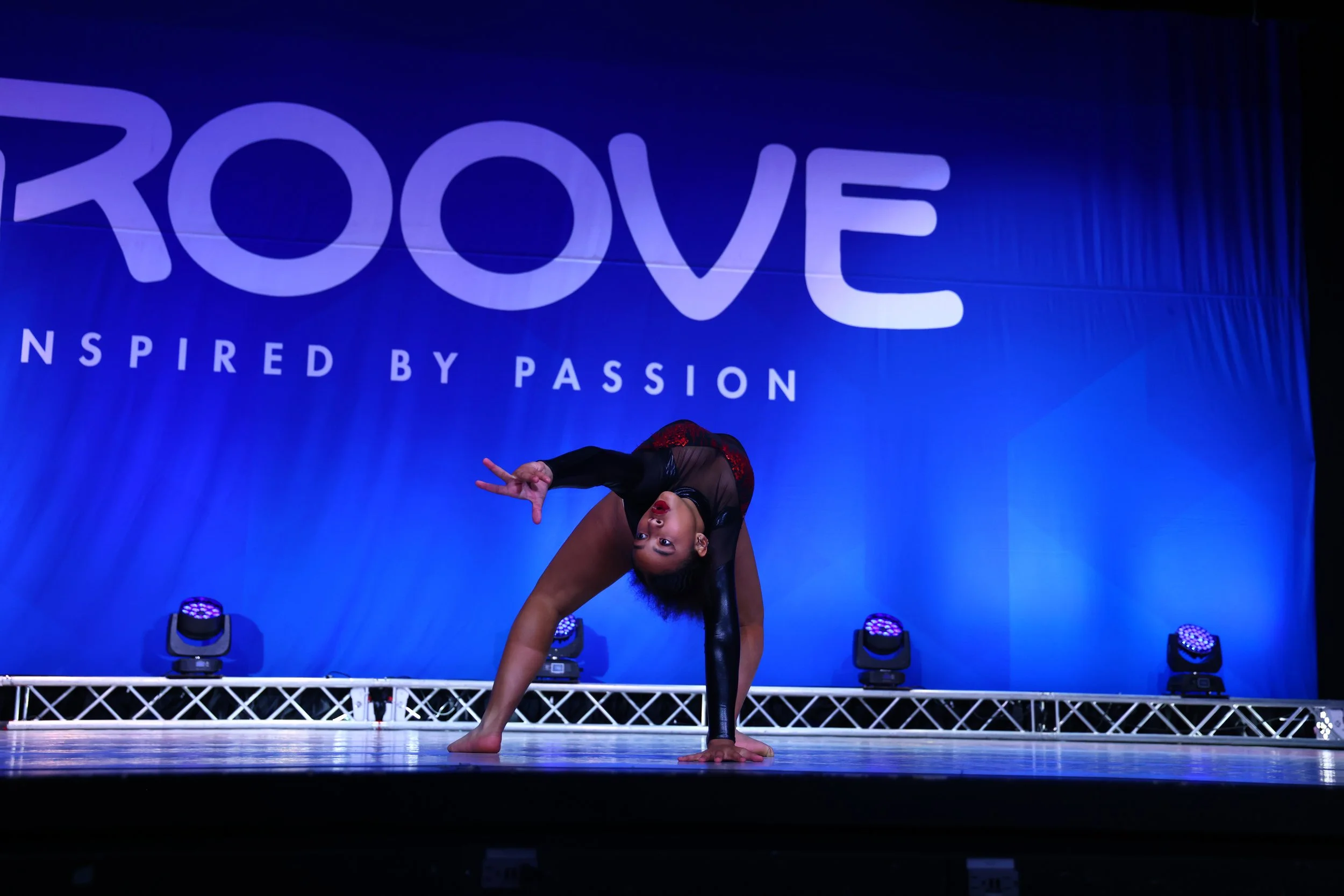 A female gymnast performing a pose on stage during a competition with a blue backdrop that says 'GROOVE' and 'INSPIRED BY PASSION'.