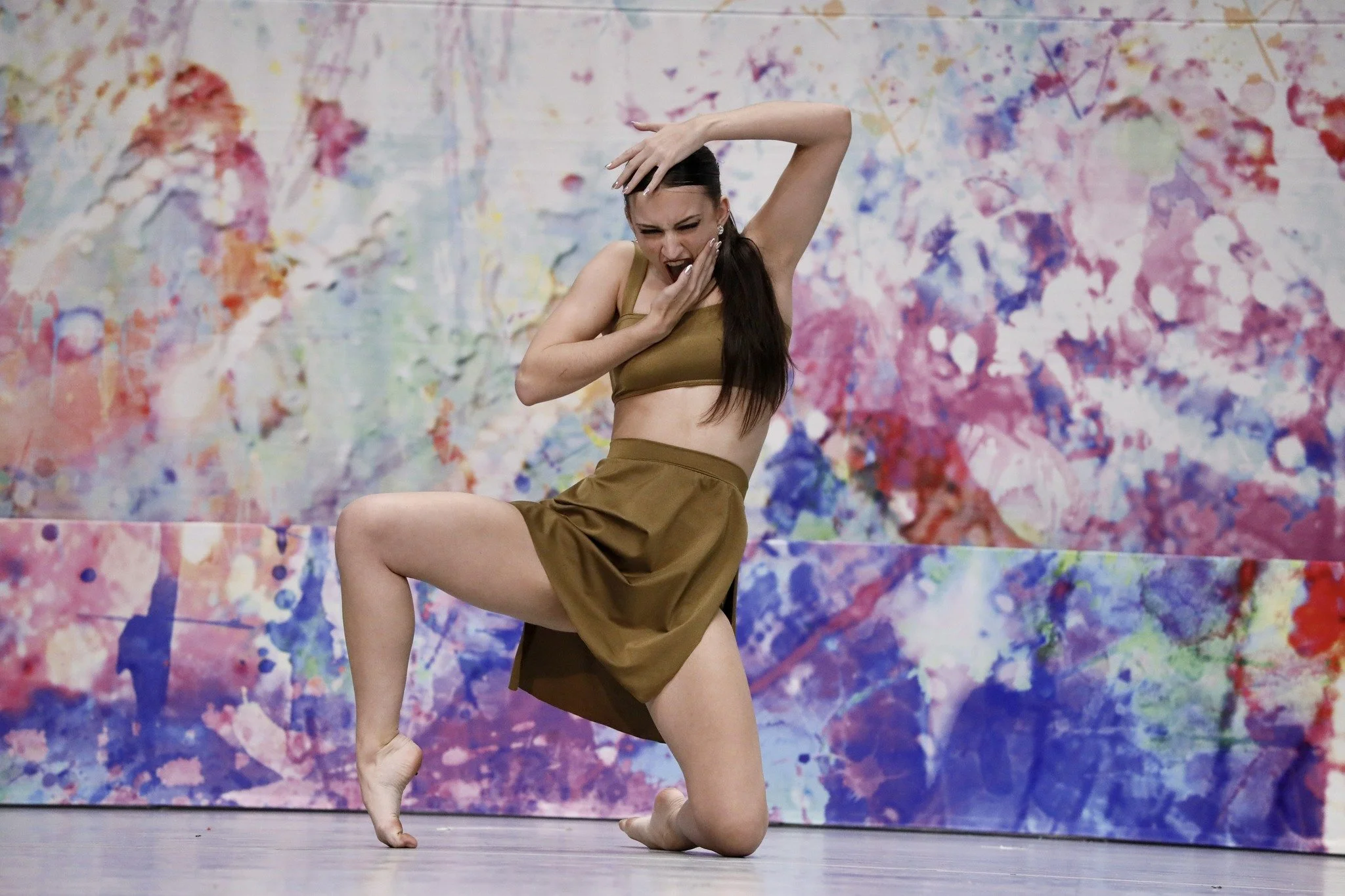 A woman dancing in front of a colorful abstract background, wearing a brown crop top and skirt, with one knee on the ground and one leg bent, expressing emotion.