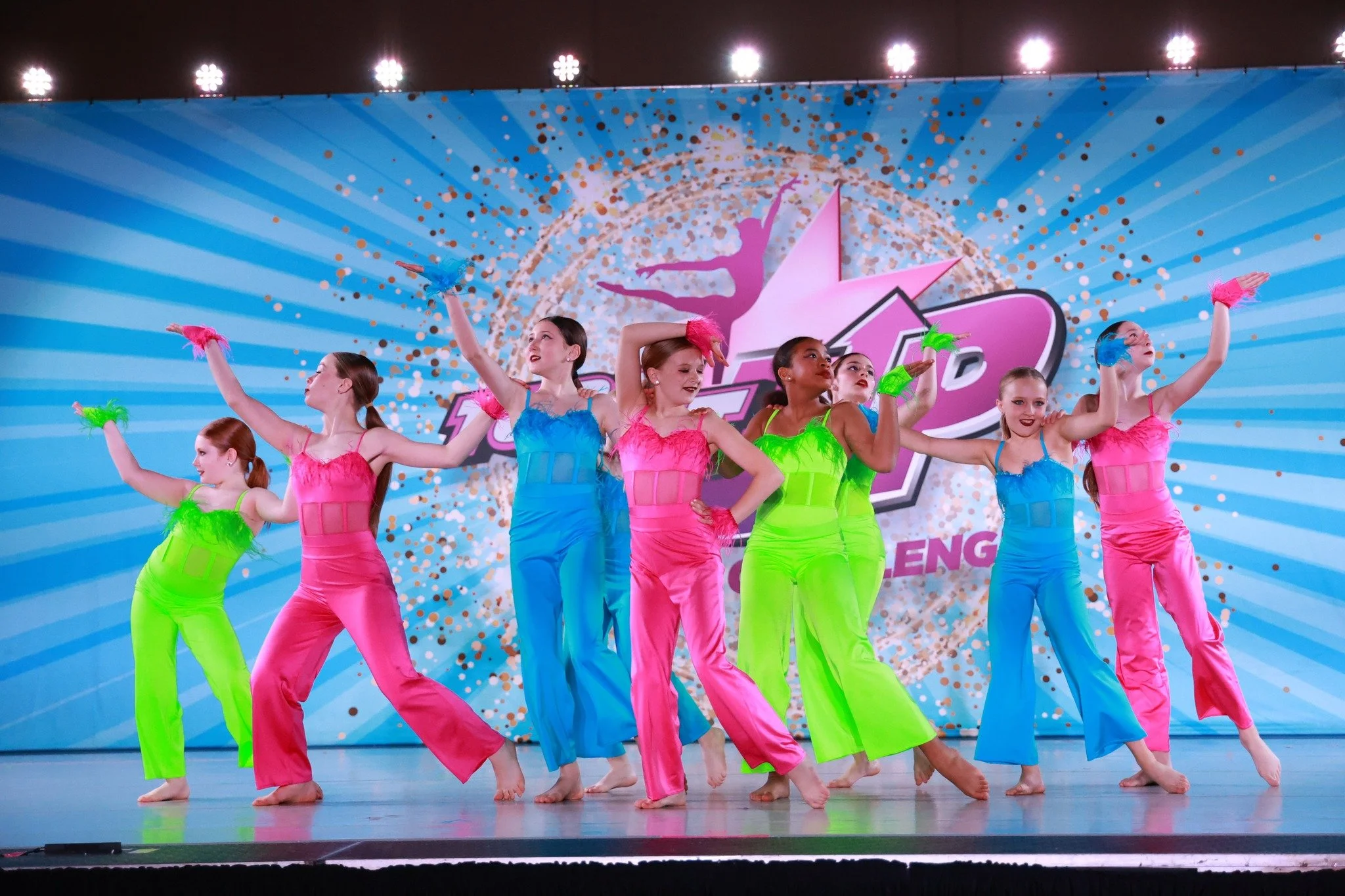 Group of young girls performing a dance on stage in colorful outfits at a dance competition or recital.