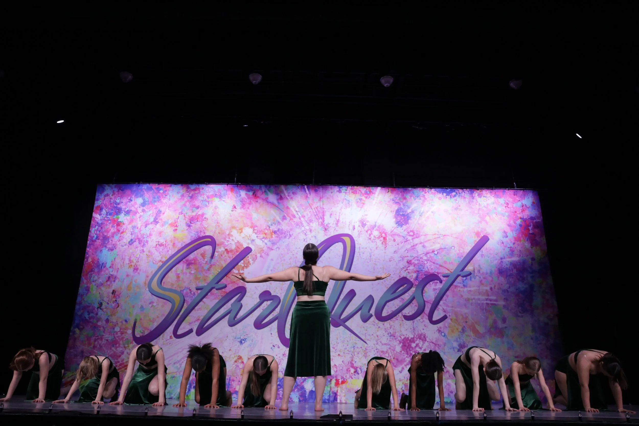 Dance performance on stage with a backdrop reading 'Startweek' in purple. One dancer in the center with back to the audience, arms outstretched, wearing a dark green outfit. Several other dancers kneeling on the stage, also in dark green costumes, bo
