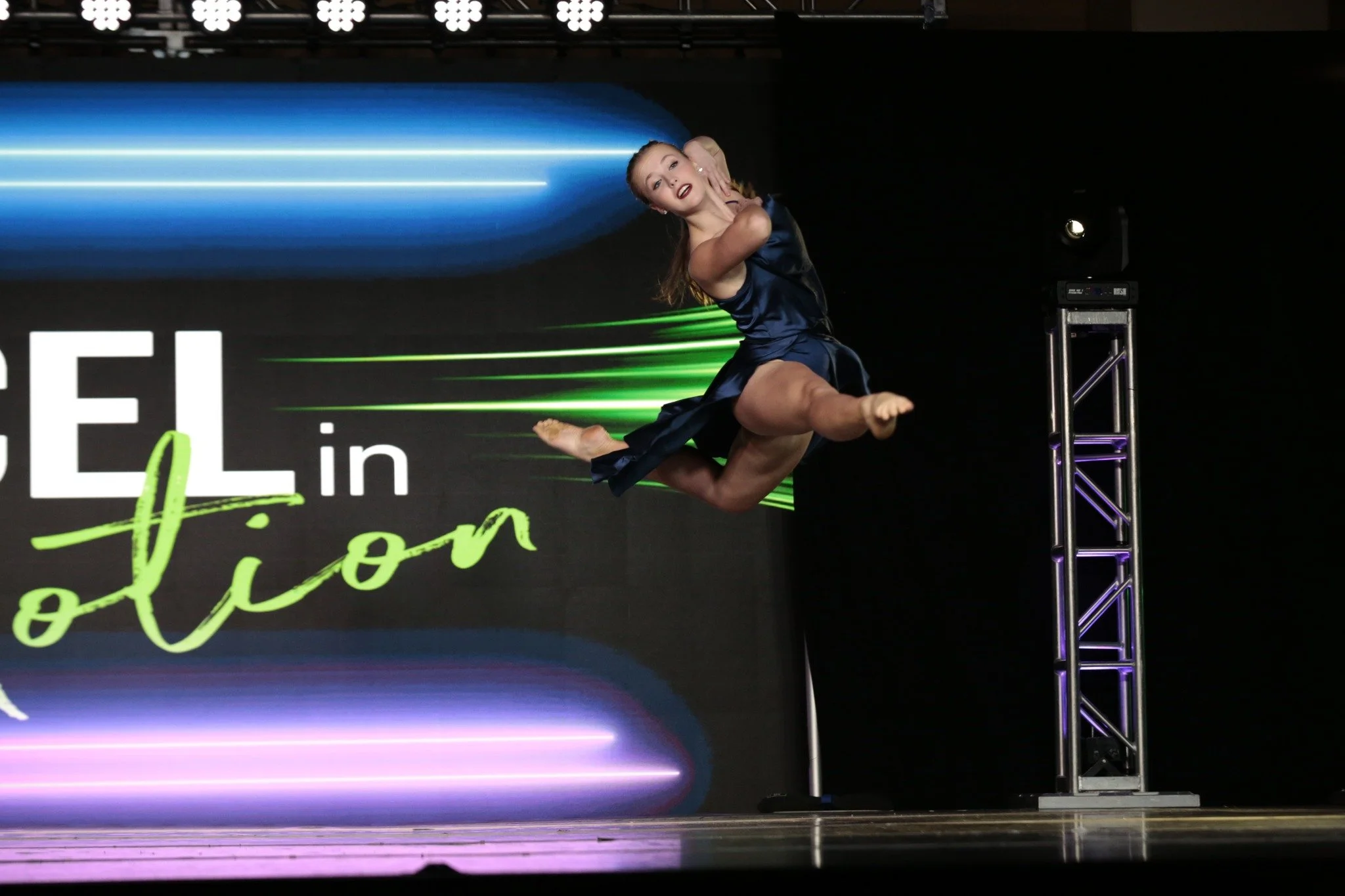 A female dancer in a dark blue dress performing a leap on stage with a black background and colorful LED lights.