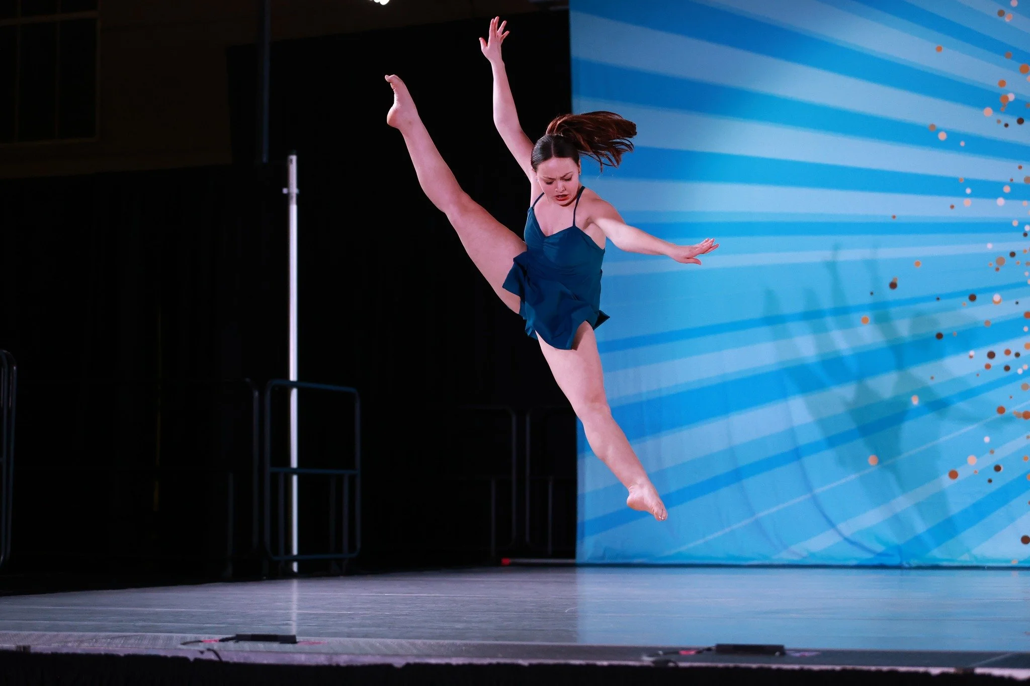 A female dancer in a blue dress performing a leap on stage, with a blue background featuring diagonal lines and orange dots.